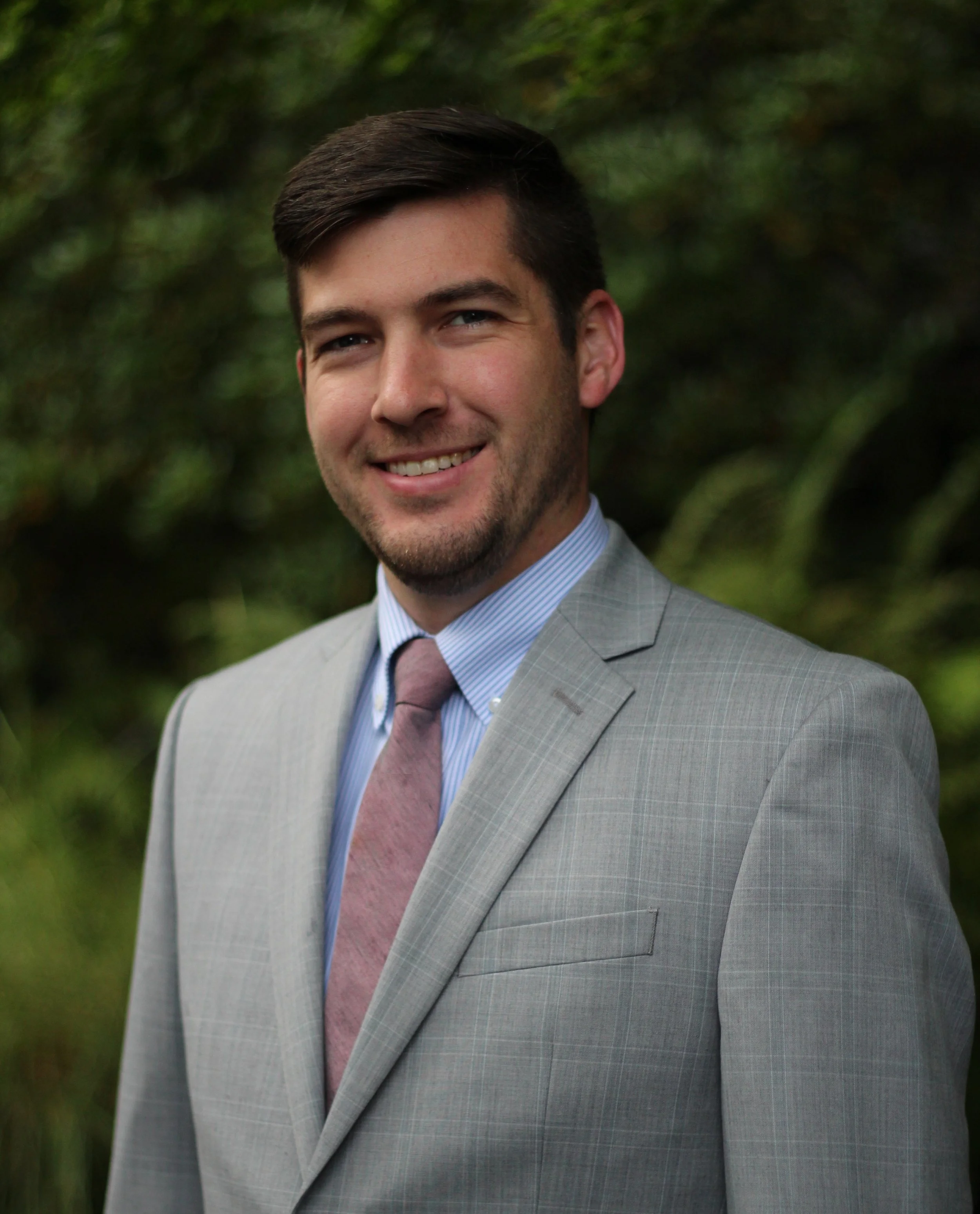 Close-up of Dr. Bryan Greenaway, an audiologist at Sono Audiology, in a gray suit, blue striped shirt, and pink tie, standing outdoors with green foliage in the background.