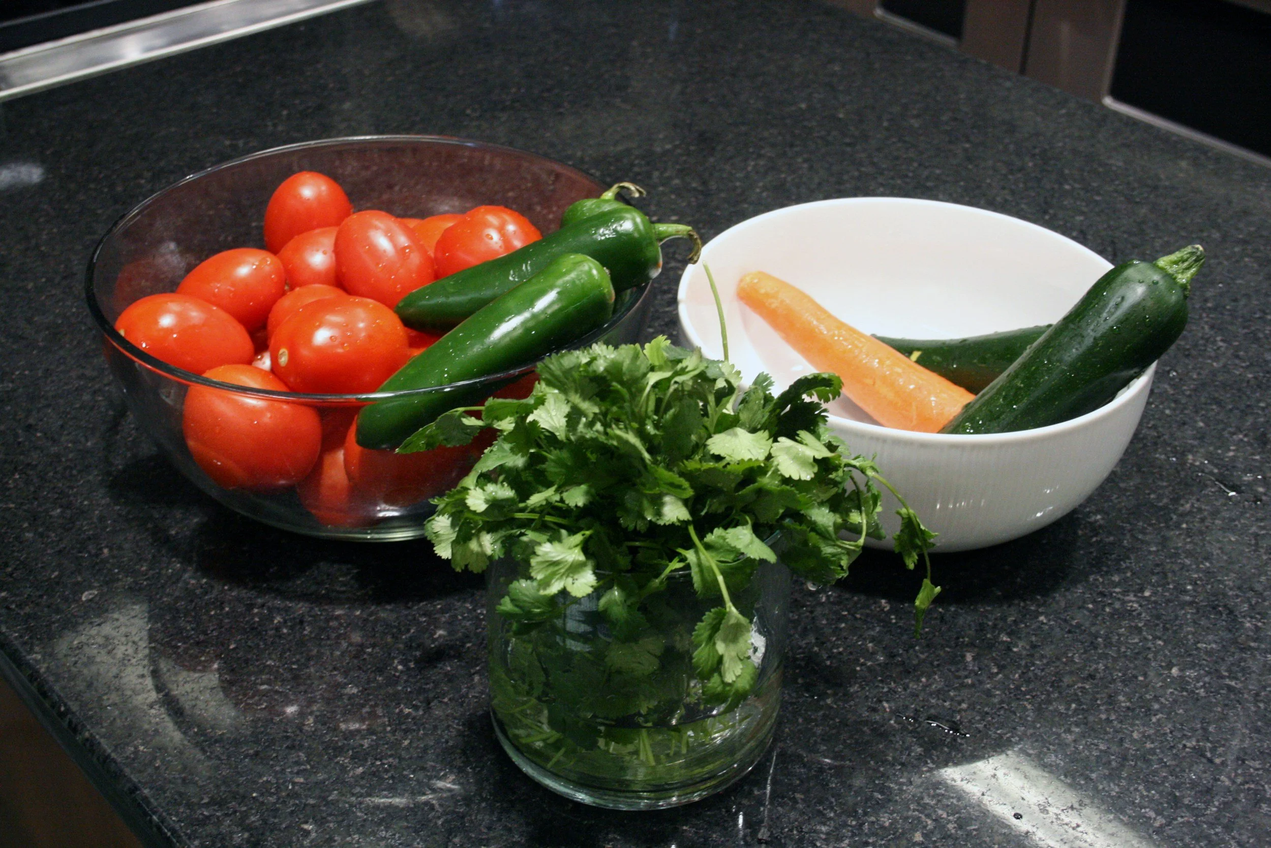 A kitchen countertop with bowls of fresh vegetables, including tomatoes, jalapeño peppers, a carrot, and cilantro.