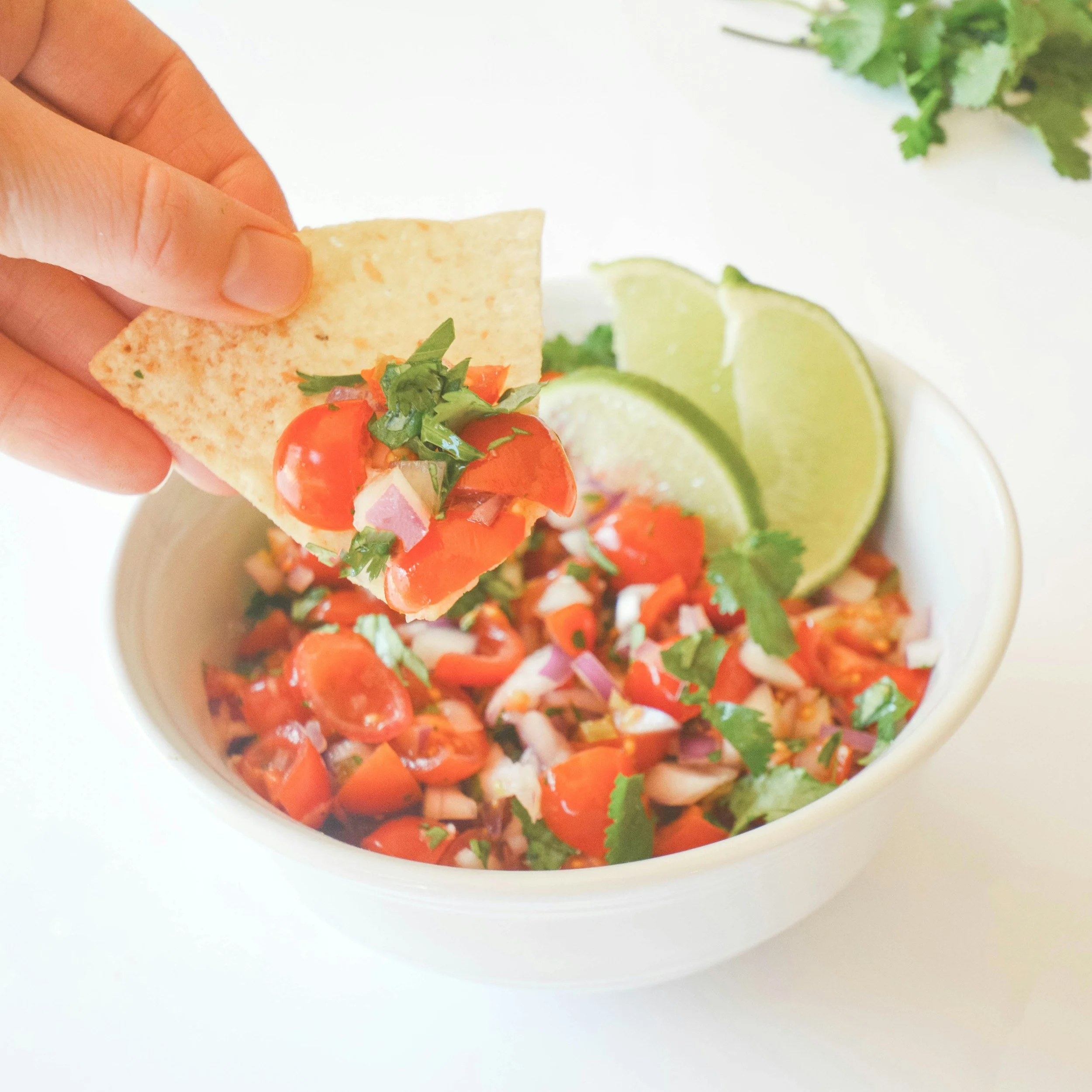 Close-up of a tortilla chip topped with chopped red tomatoes, red onions, and green cilantro, with lime wedges and green herbs in the background.