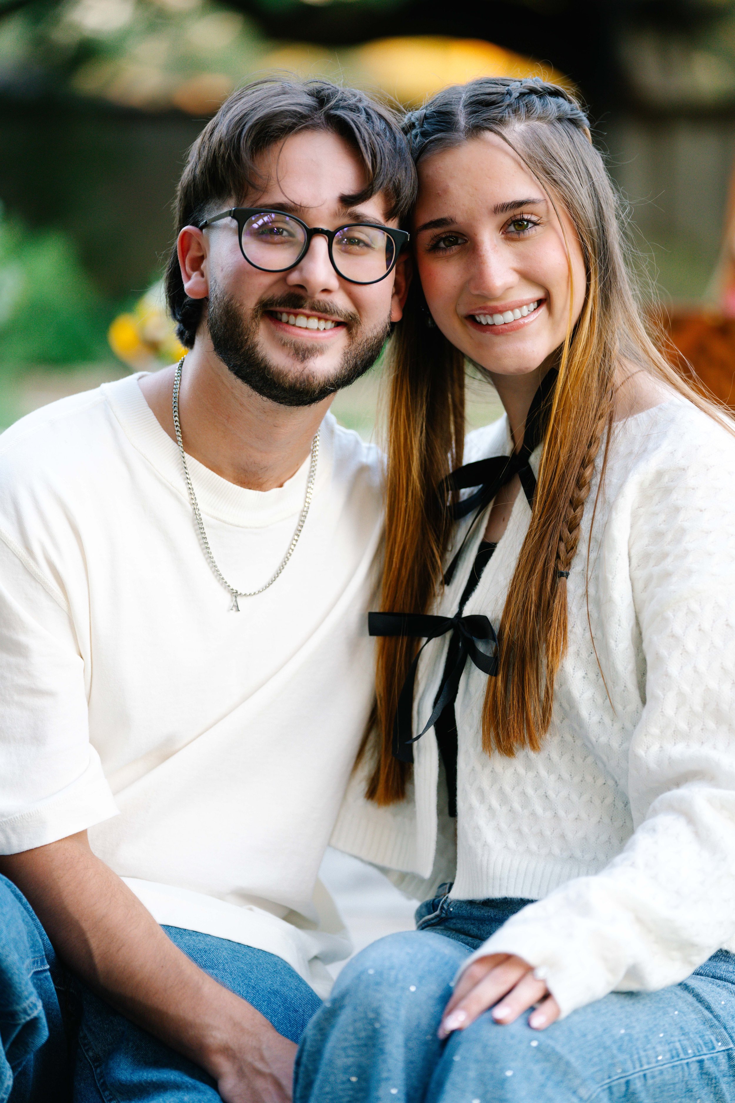Close-up of a young man with glasses and a young woman with braided hair smiling, sitting outdoors in a garden or park, with blurred greenery and yellow flowers in the background.