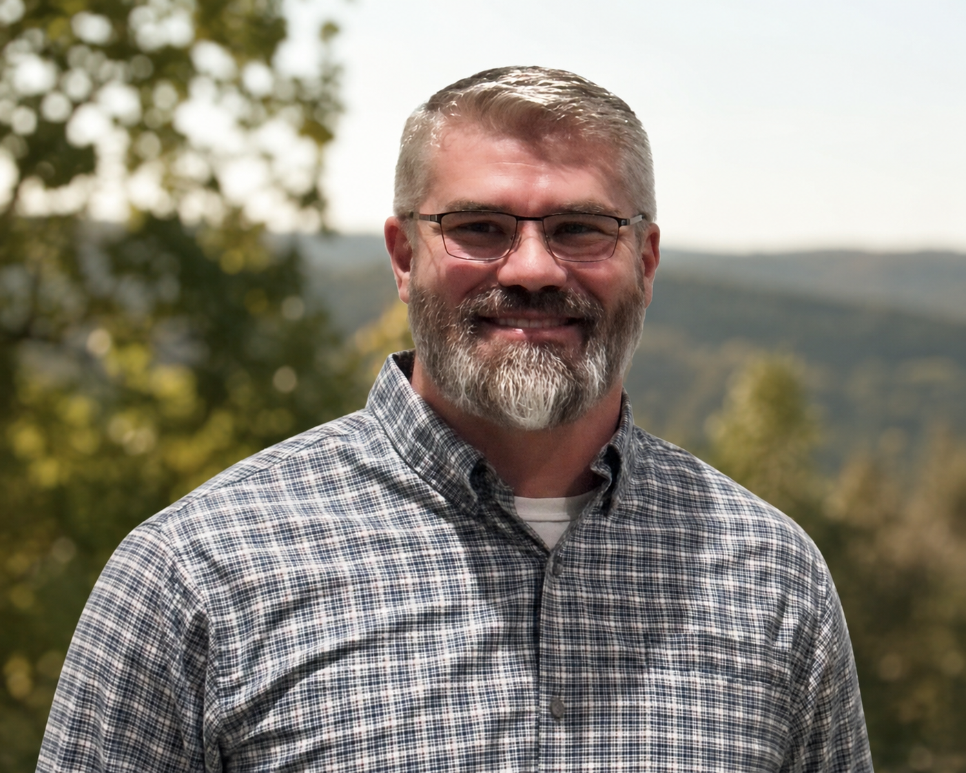 A middle-aged man with glasses, a beard, and short gray hair smiling outdoors, wearing a plaid shirt with a scenic background of trees and hills.