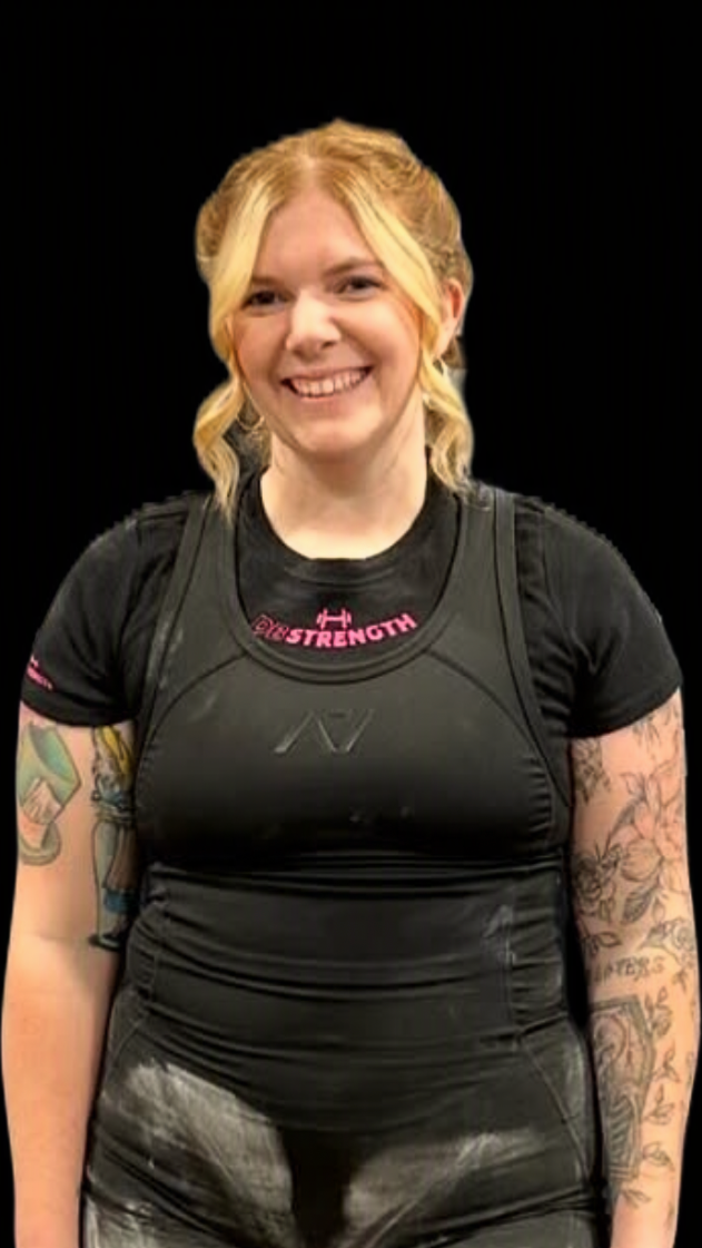 Young woman with blonde hair styled in loose curls, smiling, wearing black athletic gear with a pink hashtag and icons, tattoos on both arms, standing against a black background.