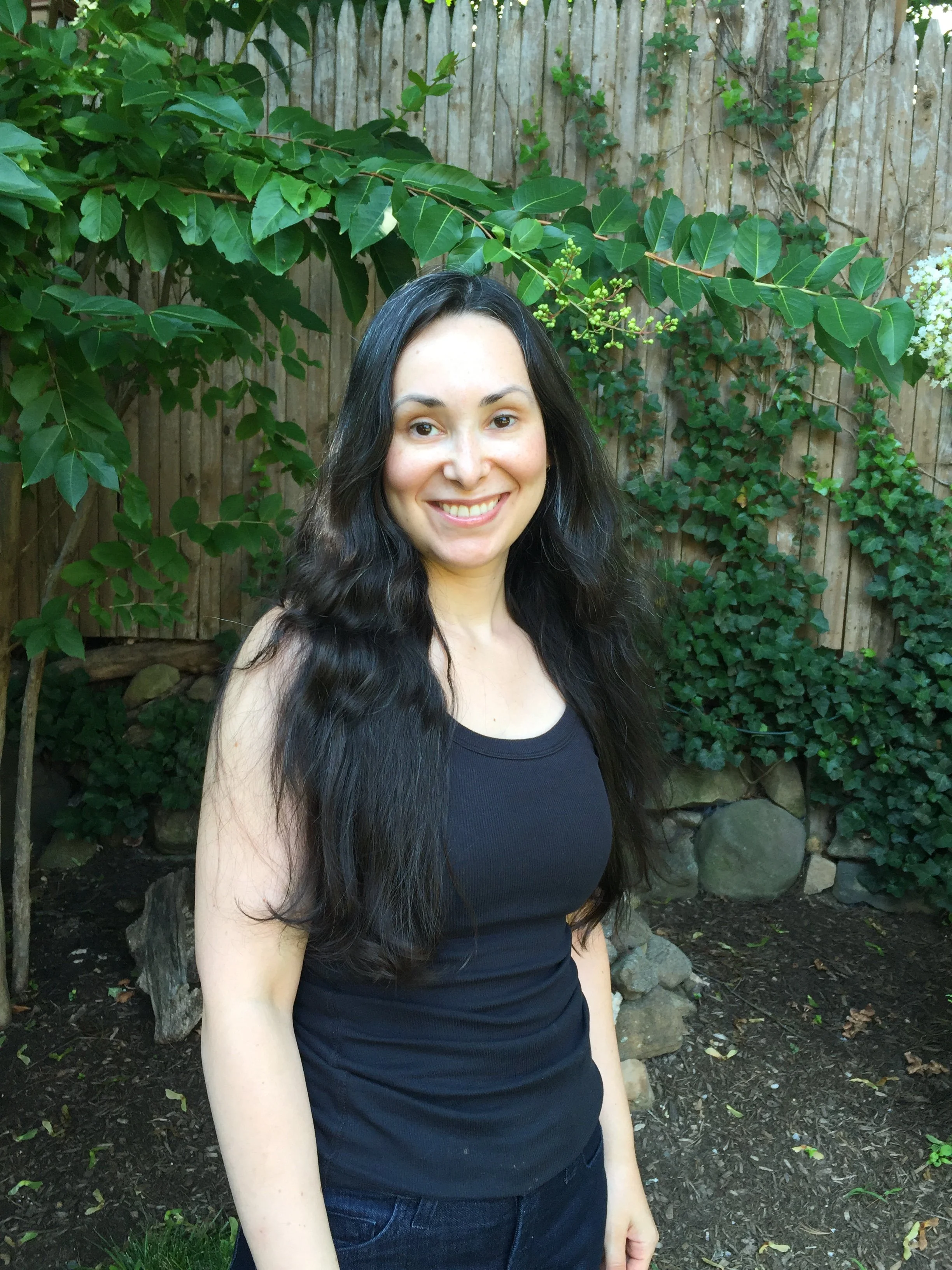 A woman with long dark hair, wearing a black top, smiling outdoors in front of a wooden fence covered in ivy and leafy plants.