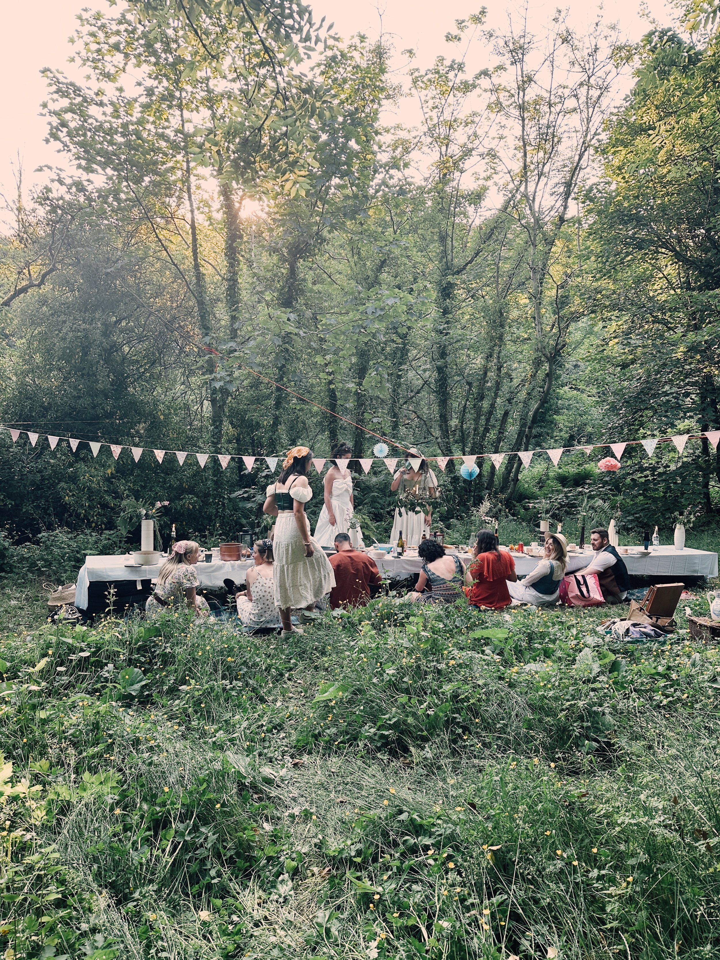 People at an outdoor gathering with a table set up, decorated with bunting and hanging paper lanterns, in a lush green forest during daytime.
