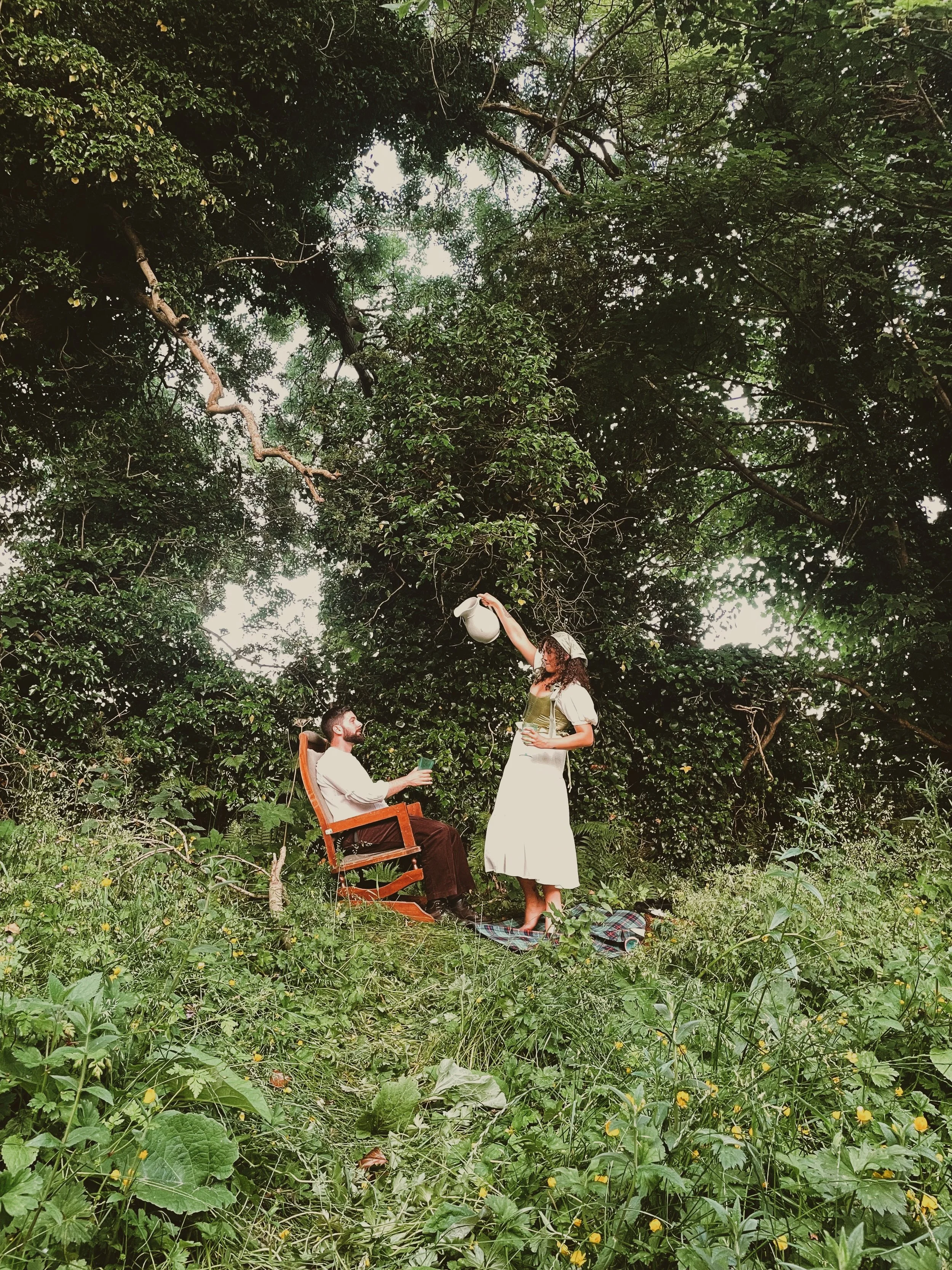 A man sitting in a wooden chair and holding a drink, while a woman standing in a white dress watering him with a watering can in a lush green outdoor setting surrounded by trees and foliage.