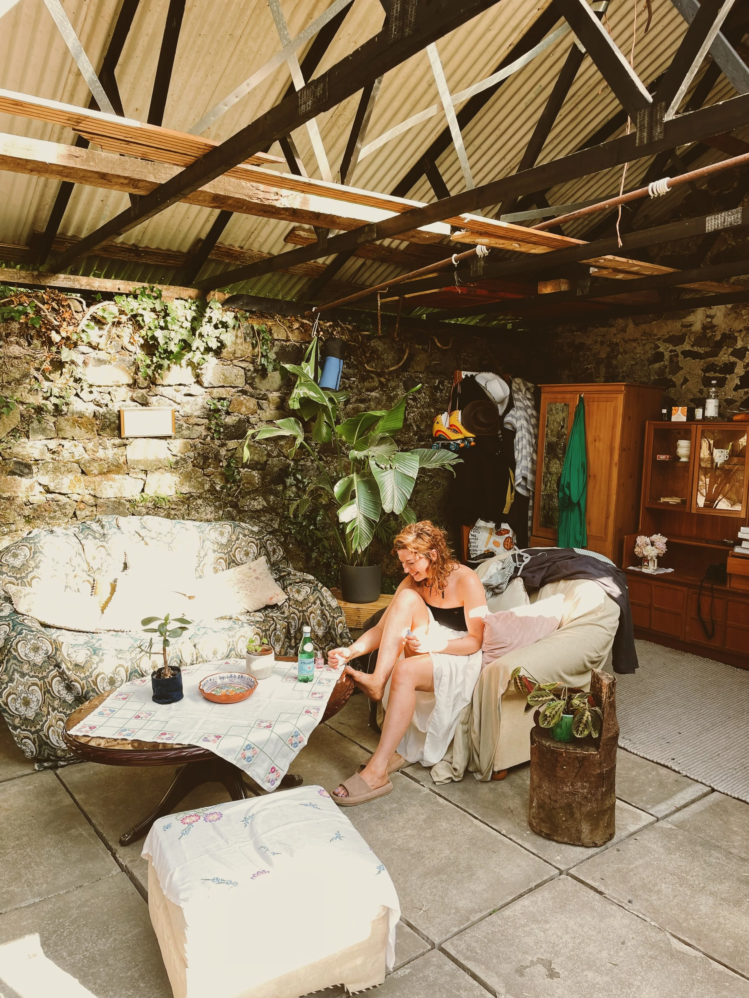 A woman sitting on a couch in a living room, bending her knees, and smiling while she appears to be putting on shoes. The room has a rustic rock wall, a large leafy plant, and a coffee table with a vase of flowers, a bottle of sparkling water, and a small plate. The ceiling has exposed wooden beams and corrugated metal roofing. There is a wooden cabinet and shelves with various items, and some clothing and accessories hanging or laying around.