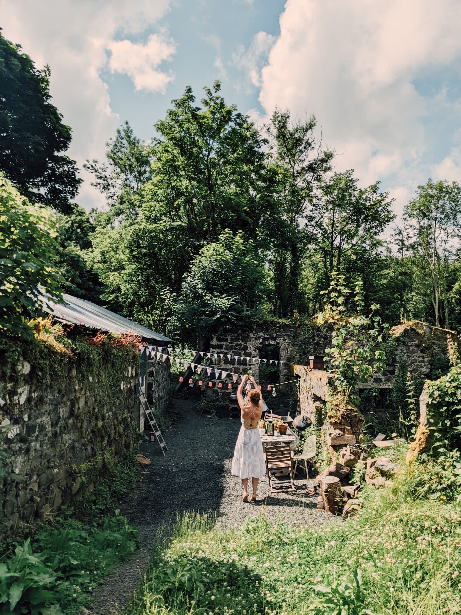 A woman in a white dress stands in a lush, green garden with trees and flowers, preparing for an outdoor event on a sunny day.