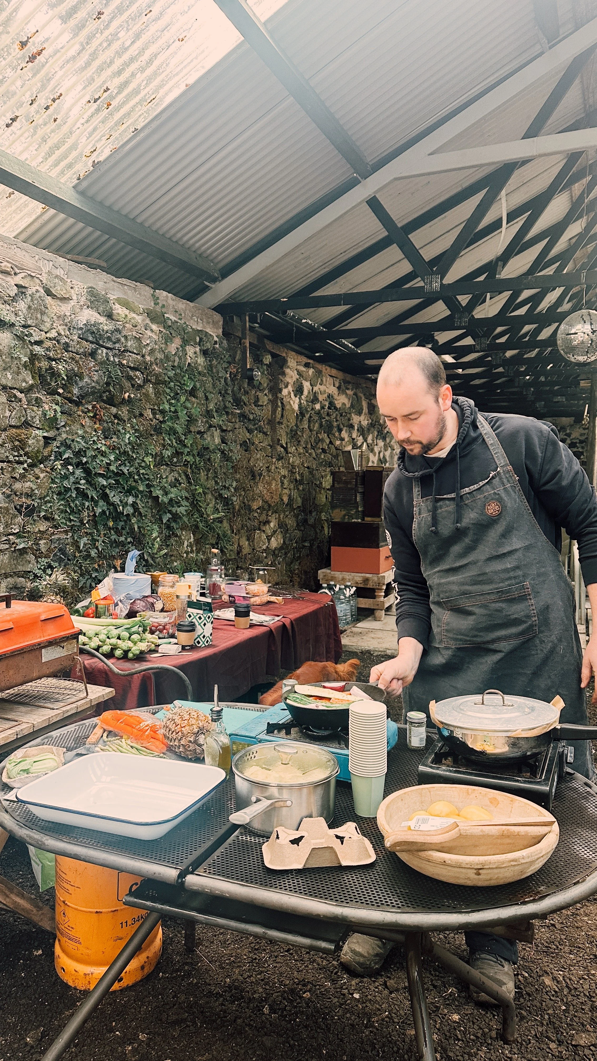 A man cooking food at an outdoor barbecue or cookout with a table of various food items and ingredients, set against a stone wall with ivy, under a metal roof.