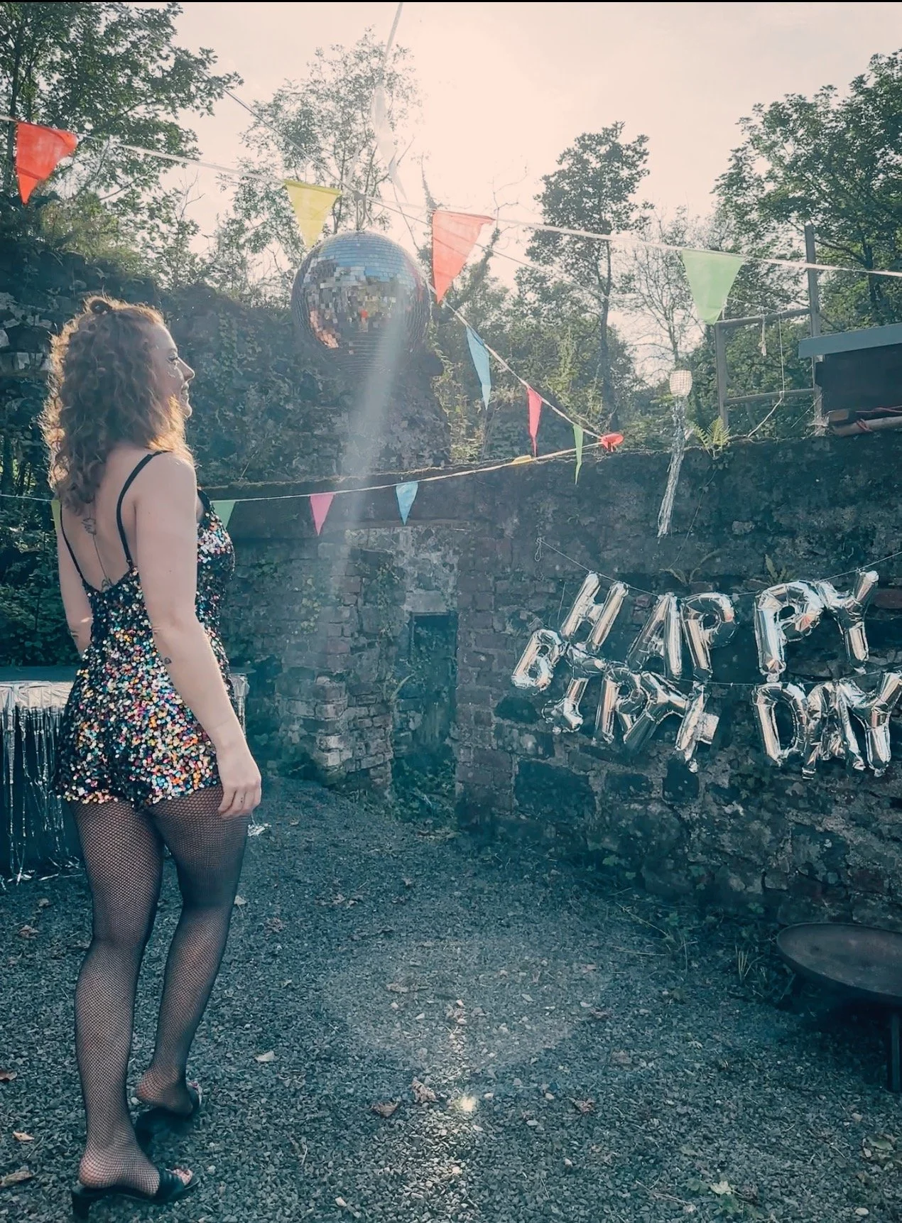 A woman with curly hair wearing a sparkly dress and fishnet stockings standing outdoors near a brick wall decorated with colorful flags and a 'Happy Birthday' banner. There is a large disco ball hanging above her.
