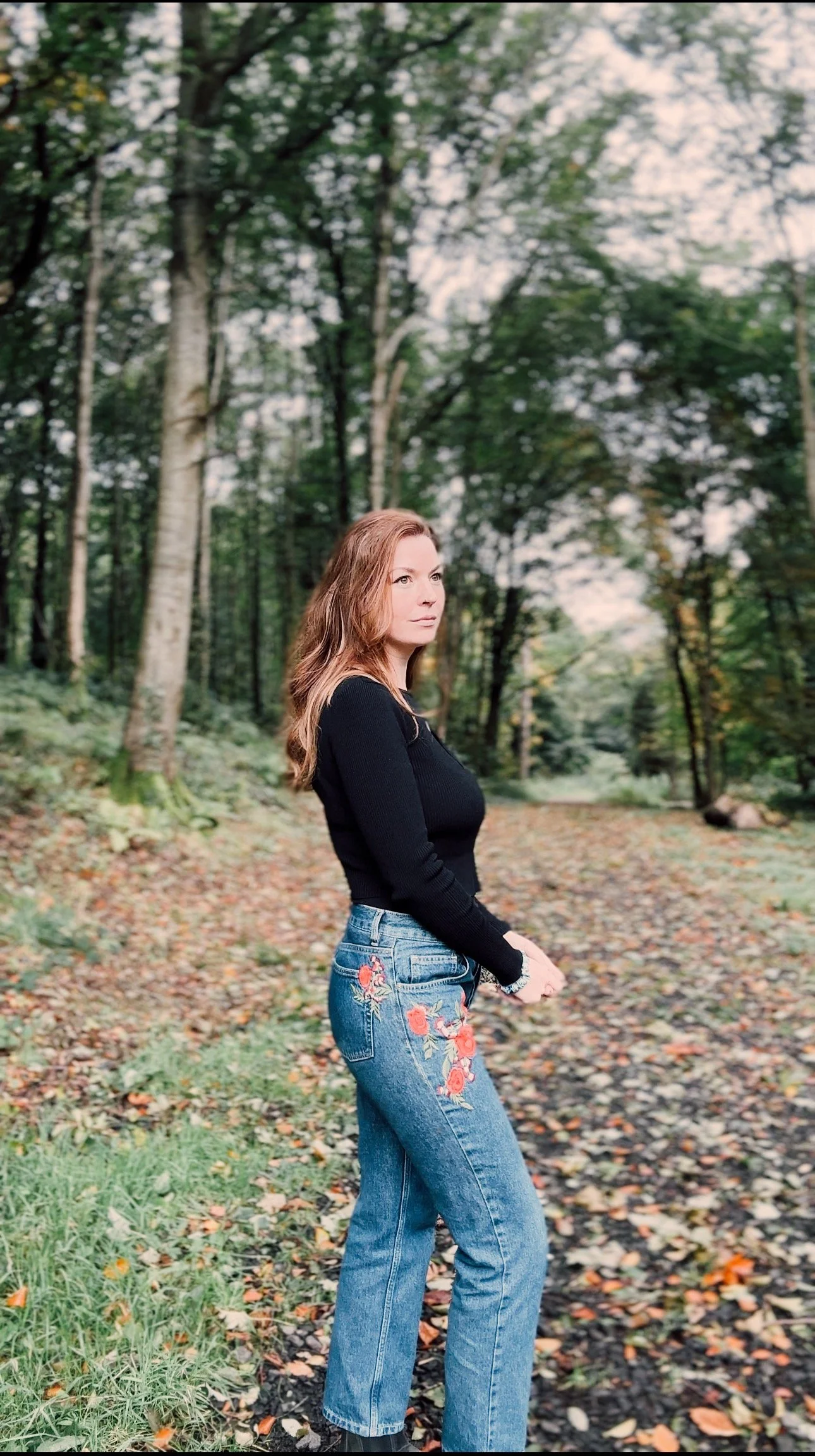 A woman with long wavy hair standing in a forest with trees and fallen leaves, wearing a black long-sleeved top and blue jeans with floral embroidery.