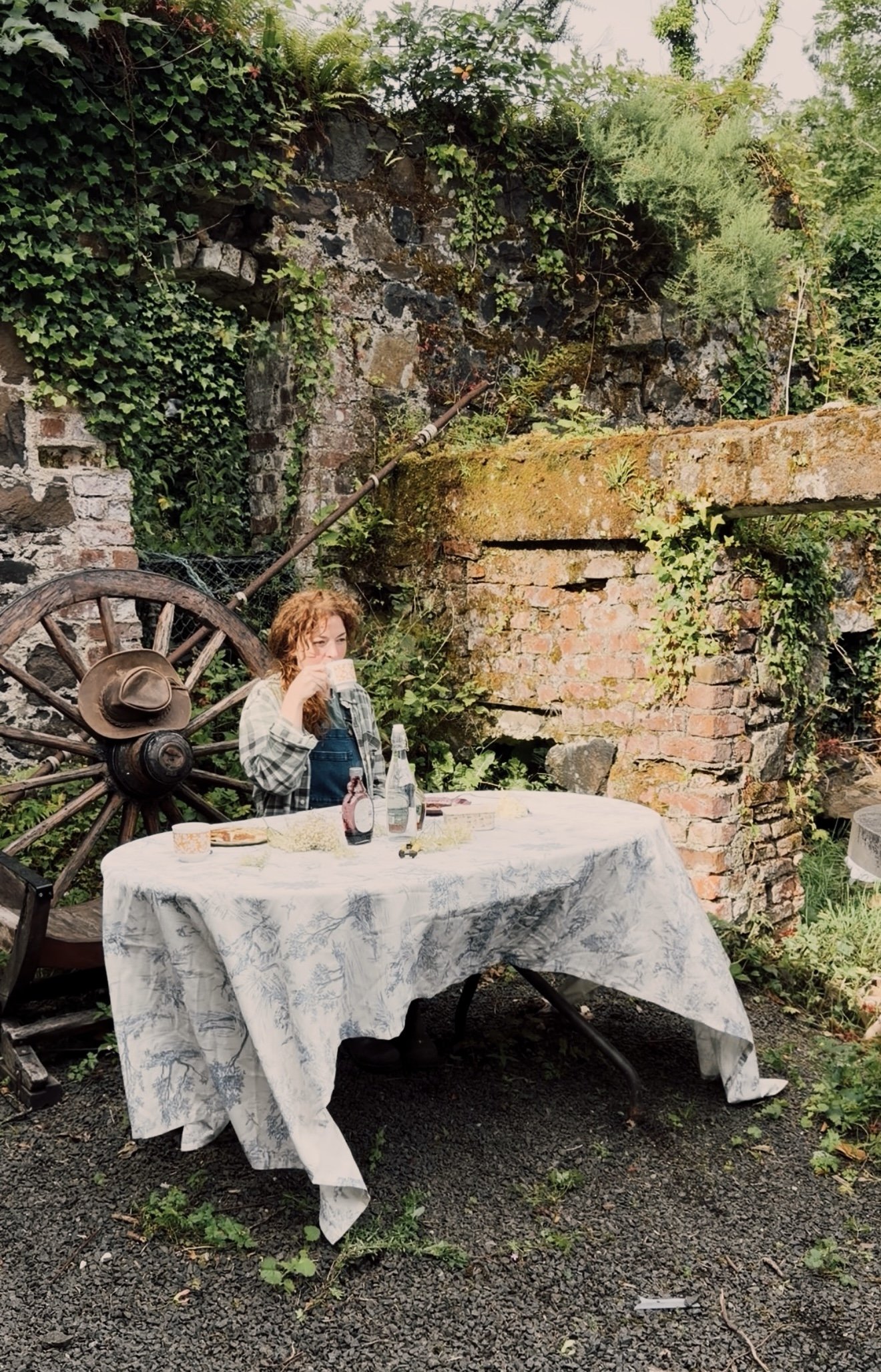 A woman with curly red hair drinking from a glass at an outdoor table covered with a white cloth, set against a rustic brick and stone wall with green ivy and plants, and a large wooden wagon wheel leaning against the wall.