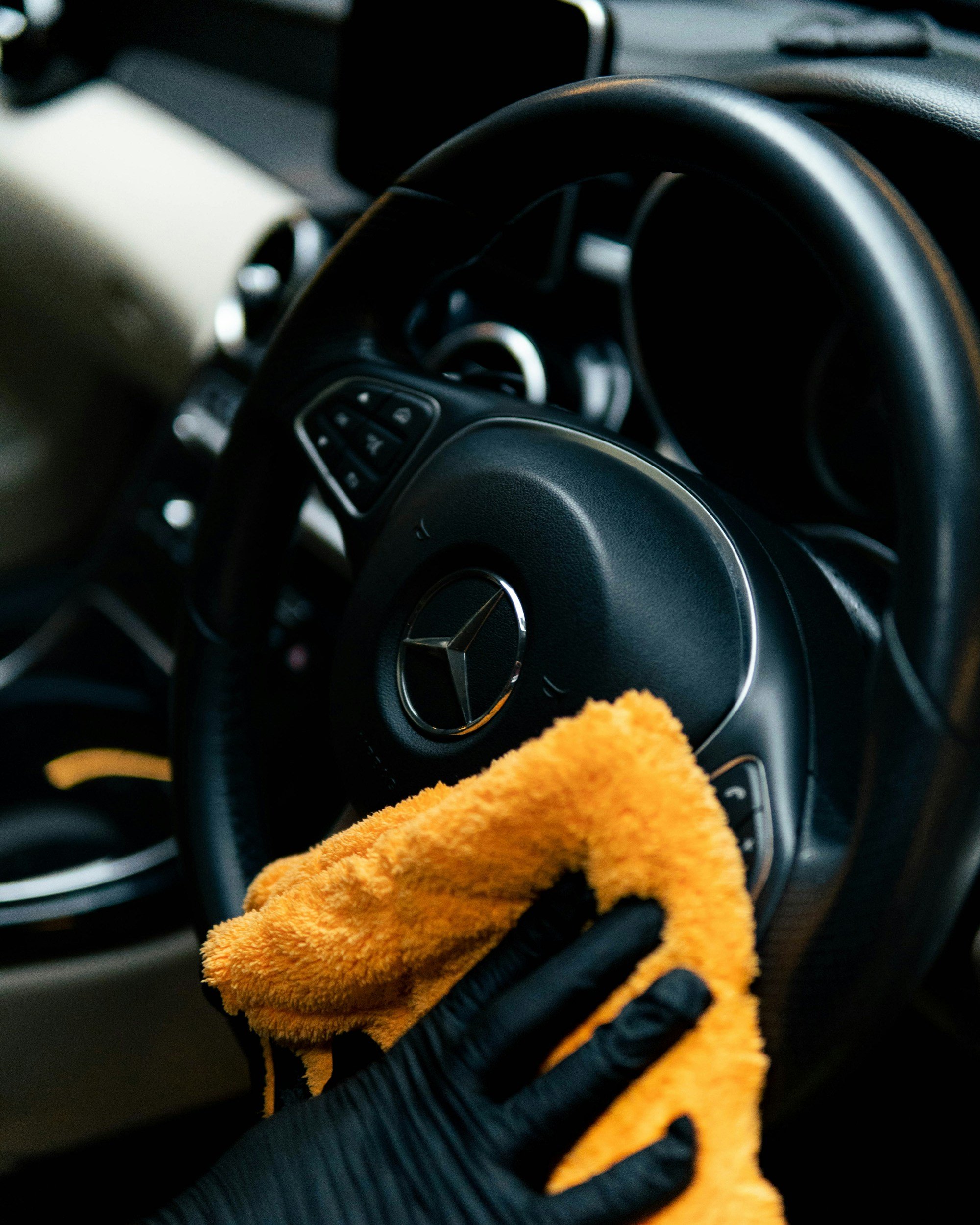 Close-up of a car's steering wheel with Mercedes-Benz logo and an orange cleaning cloth in a hand, with the dashboard and air conditioning vent in the background.