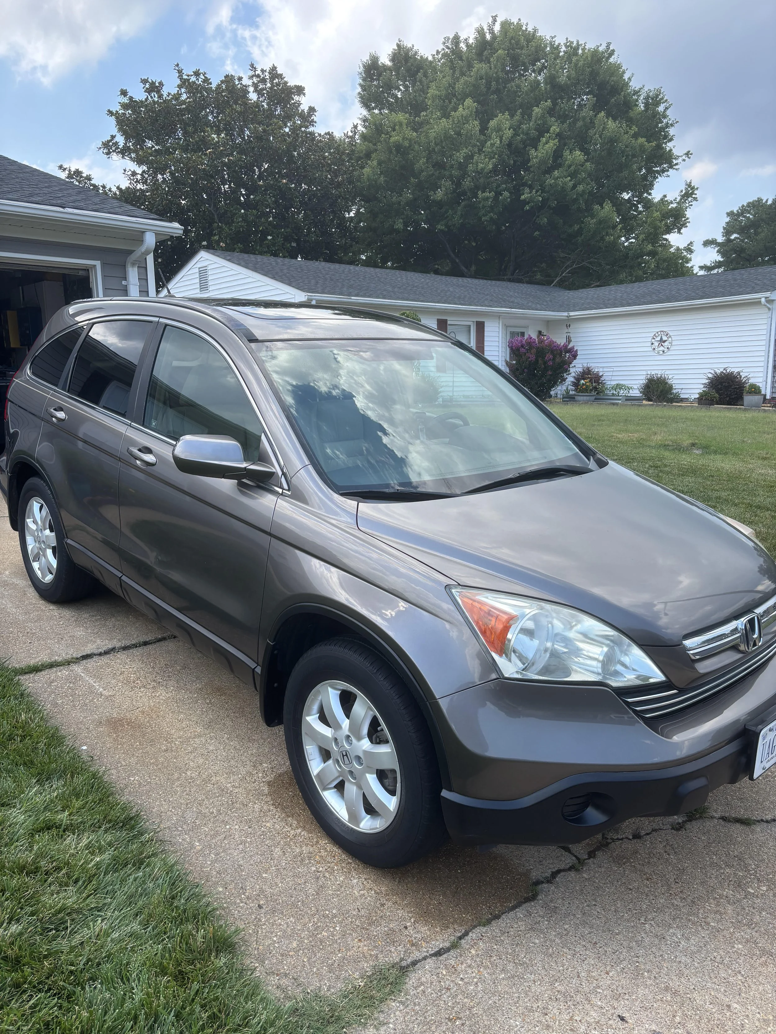 A gray Honda CR-V parked on a concrete driveway with grass on the left, in front of a suburban house with white siding, a gable roof, and a large tree in the background.