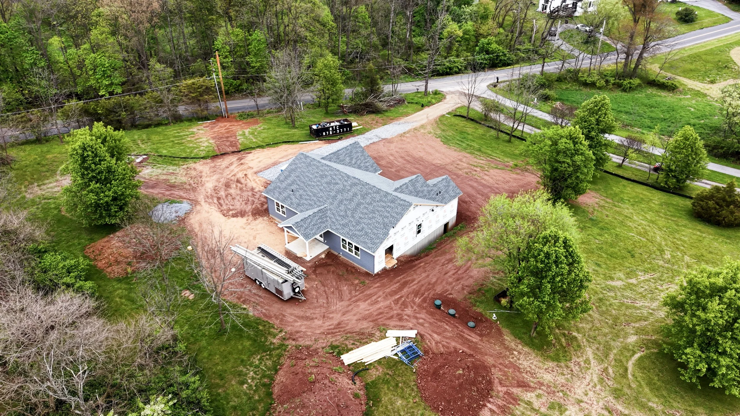 A house under construction with a gray roof, surrounded by dirt and green trees, a utility trailer and construction materials nearby, and a road in the background.