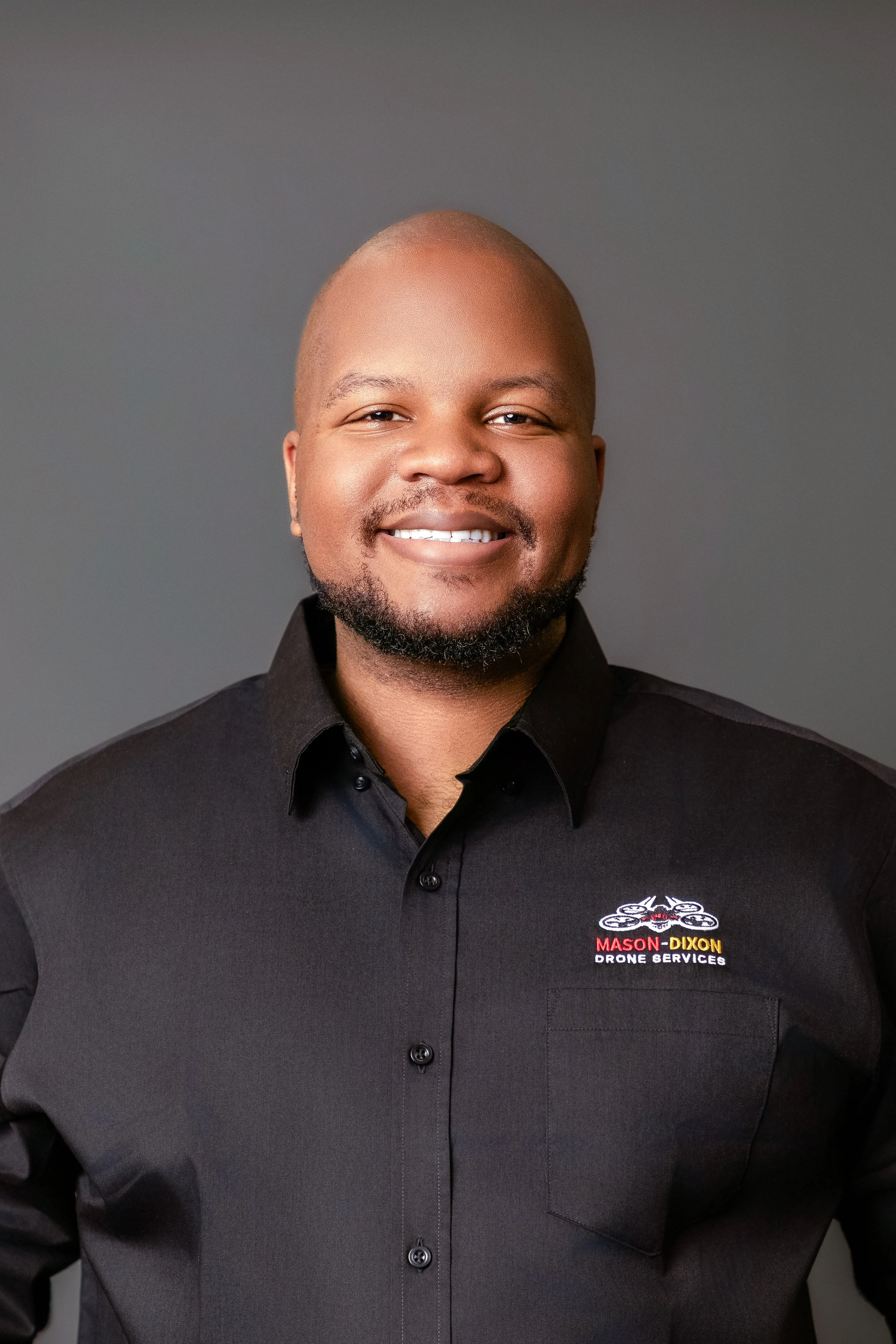 A smiling man with a bald head and dark beard, wearing a black button-up shirt with a logo for Mason-Dixon Drone Services on it. The background is plain and gray.