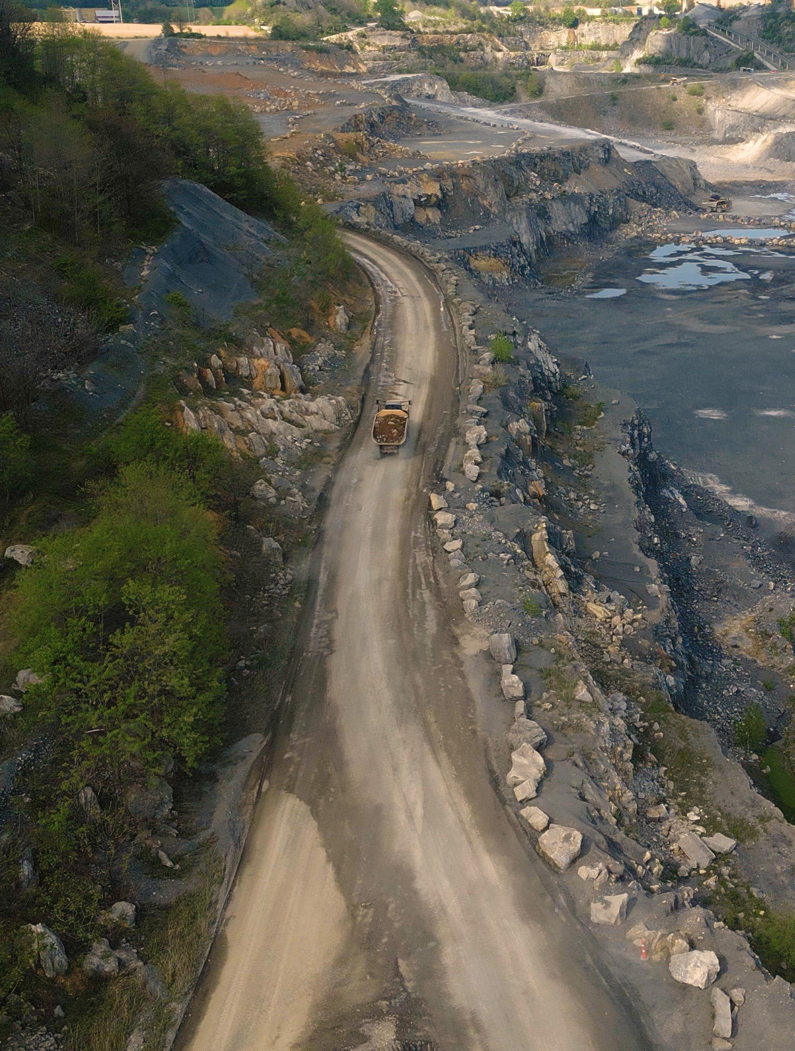 A gravel road winding through a rocky and hilly landscape, with trees on the left and a body of water on the right. Construction equipment and disturbed land are visible in the background.