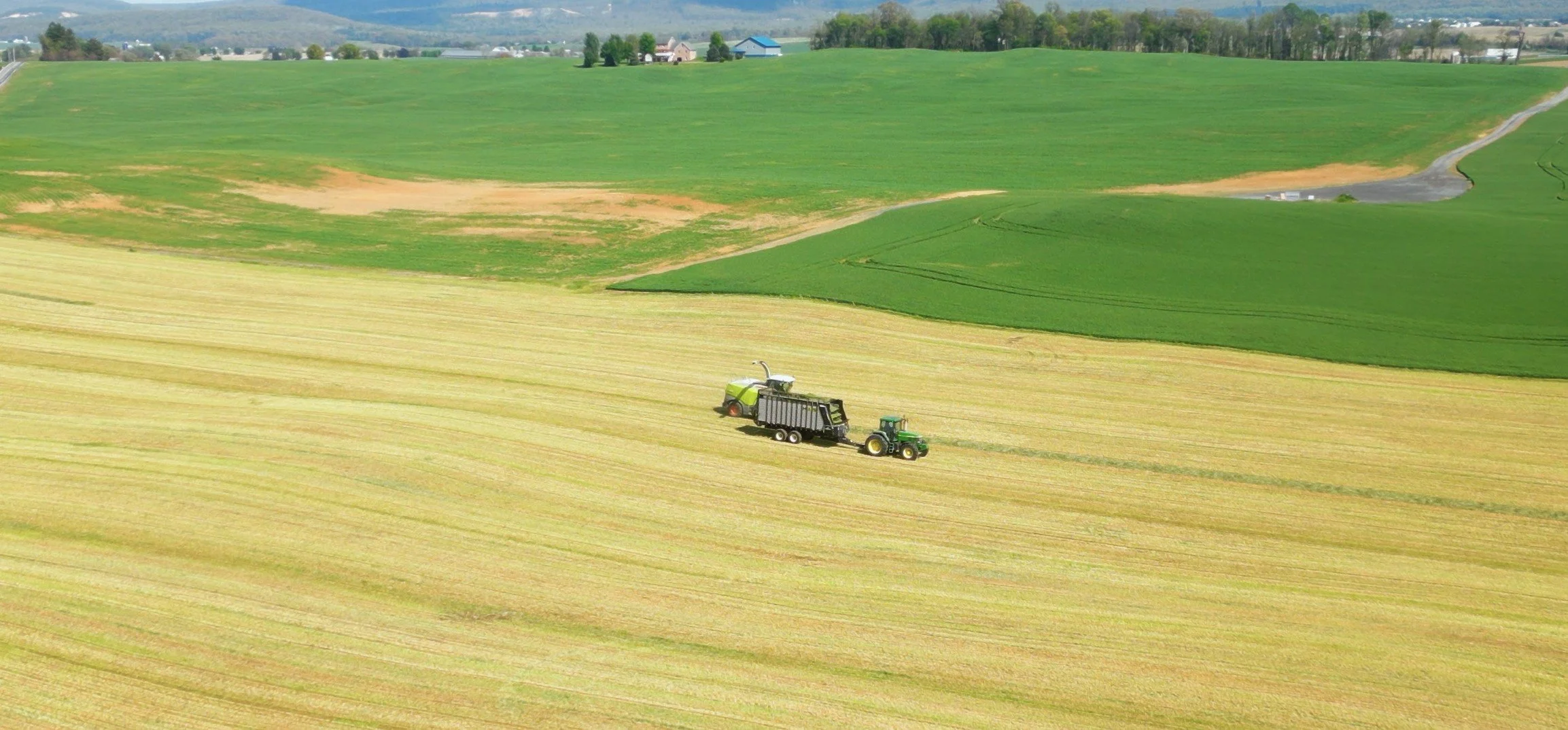 An aerial view of a green field with a tractor pulling a storage trailer in a wheat field, with rolling hills and farm buildings in the background.