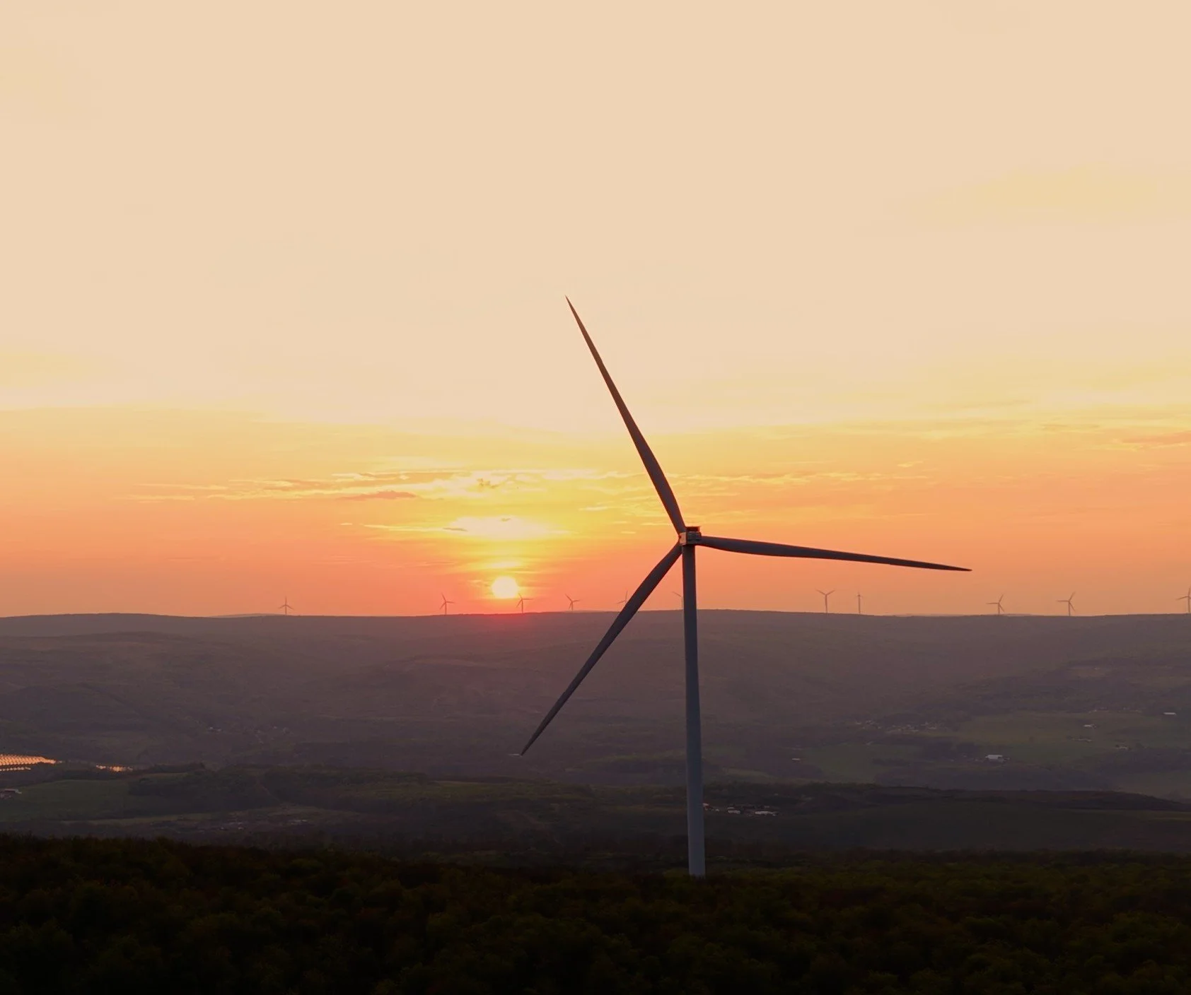A landscape with a wind turbine in the foreground during sunset, with additional wind turbines in the background, rolling hills, and a partly cloudy sky.
