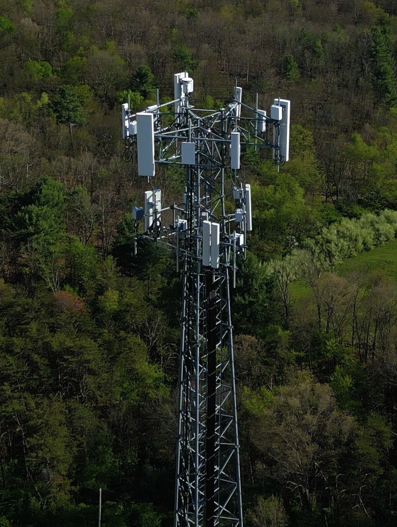 A tall telecommunications tower with multiple antennas is situated in a lush, wooded area with green trees.