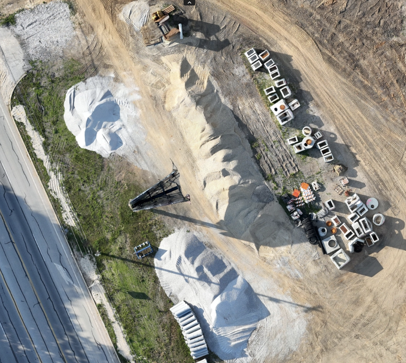 An aerial view of a quarry or construction site with large piles of gravel, a conveyor system, and materials stored in various containers. A highway runs along the side with power lines crossing the scene.