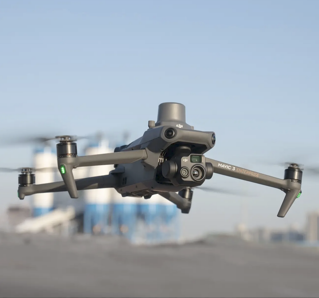 A DJI Mavic 3 Enterprise drone flying outdoors with a blurred urban background and blue sky.