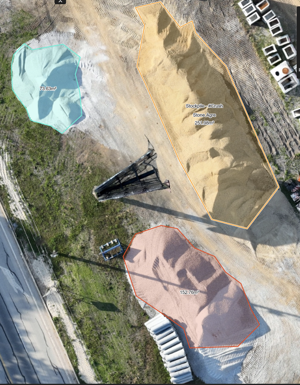 Aerial view of a construction site with piles of material, including a blue-tinted pile of gravel, a large yellow pile of stone labeled 'Stone Aggregate,' and a smaller pink pile of material. The site is outlined with different colored borders and measurements, with nearby roads and vegetation.