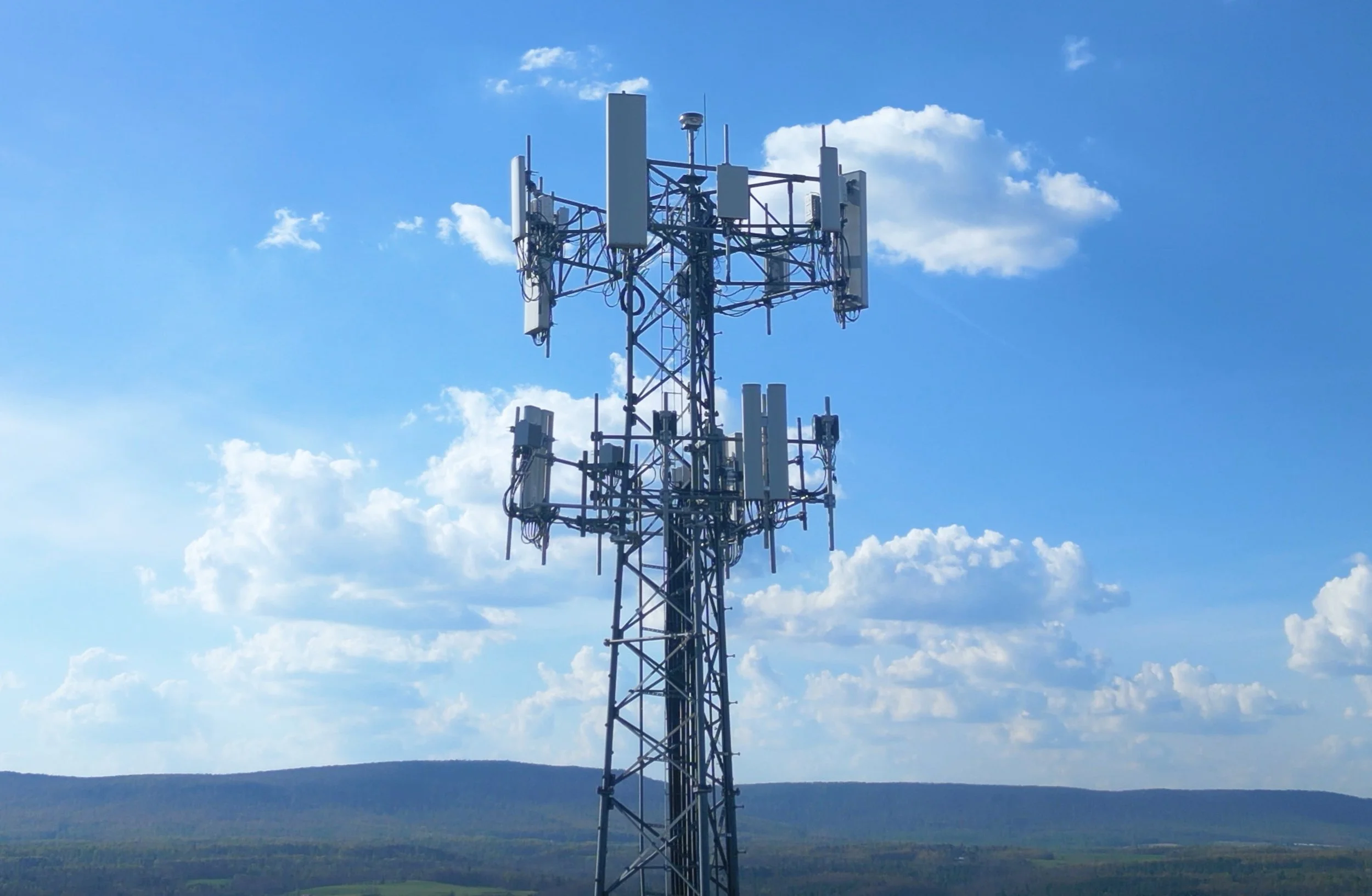 A tall communication tower with multiple antennas and dishes against a blue sky with scattered clouds and a landscape of fields and hills in the background.