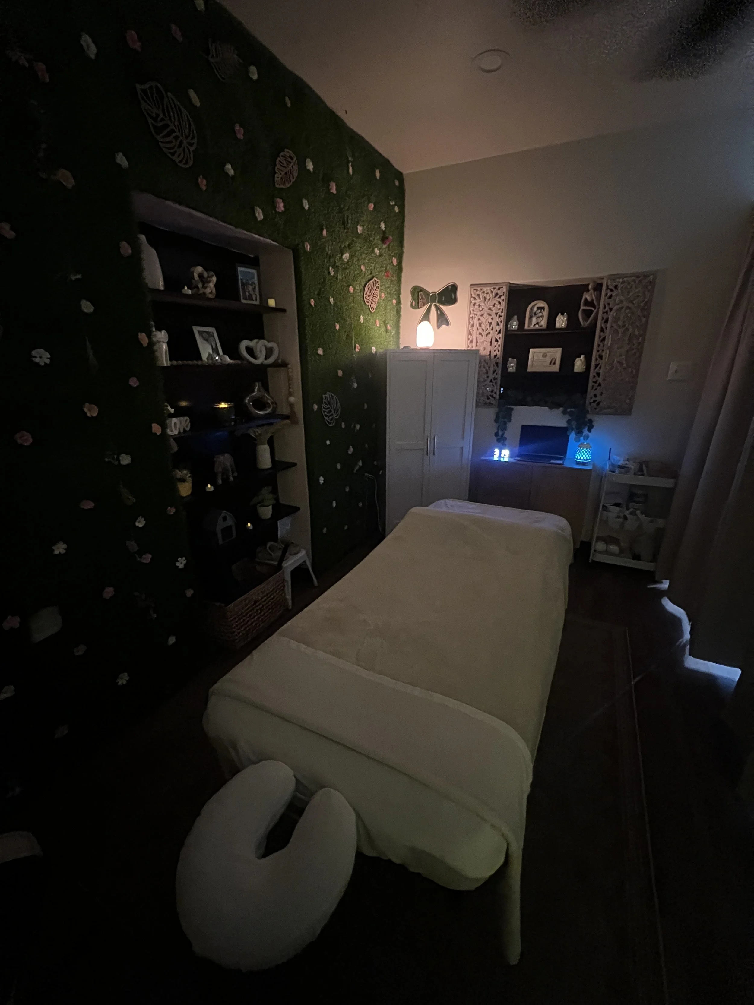 A bedroom with a bed and a clawfoot tub, decorated with a green floral wall covering and complementing shelving with various decor items and picture frames, dimly lit by a night light and a salt lamp.