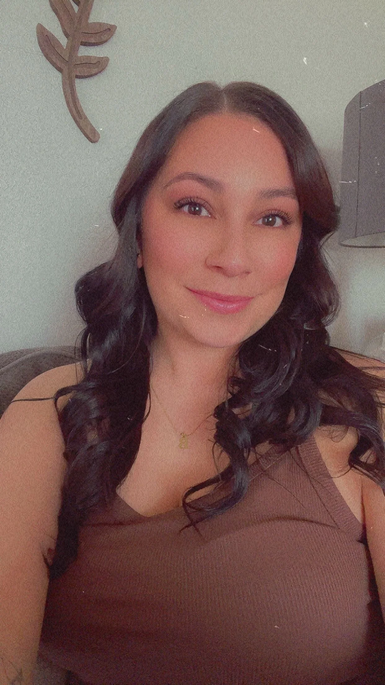 A woman with dark, curly hair is smiling and taking a selfie in a room with light-colored walls and a wall decoration of a leafless branch mounted on the wall. Owner of Breeze Massage, Brianna Gonzales.