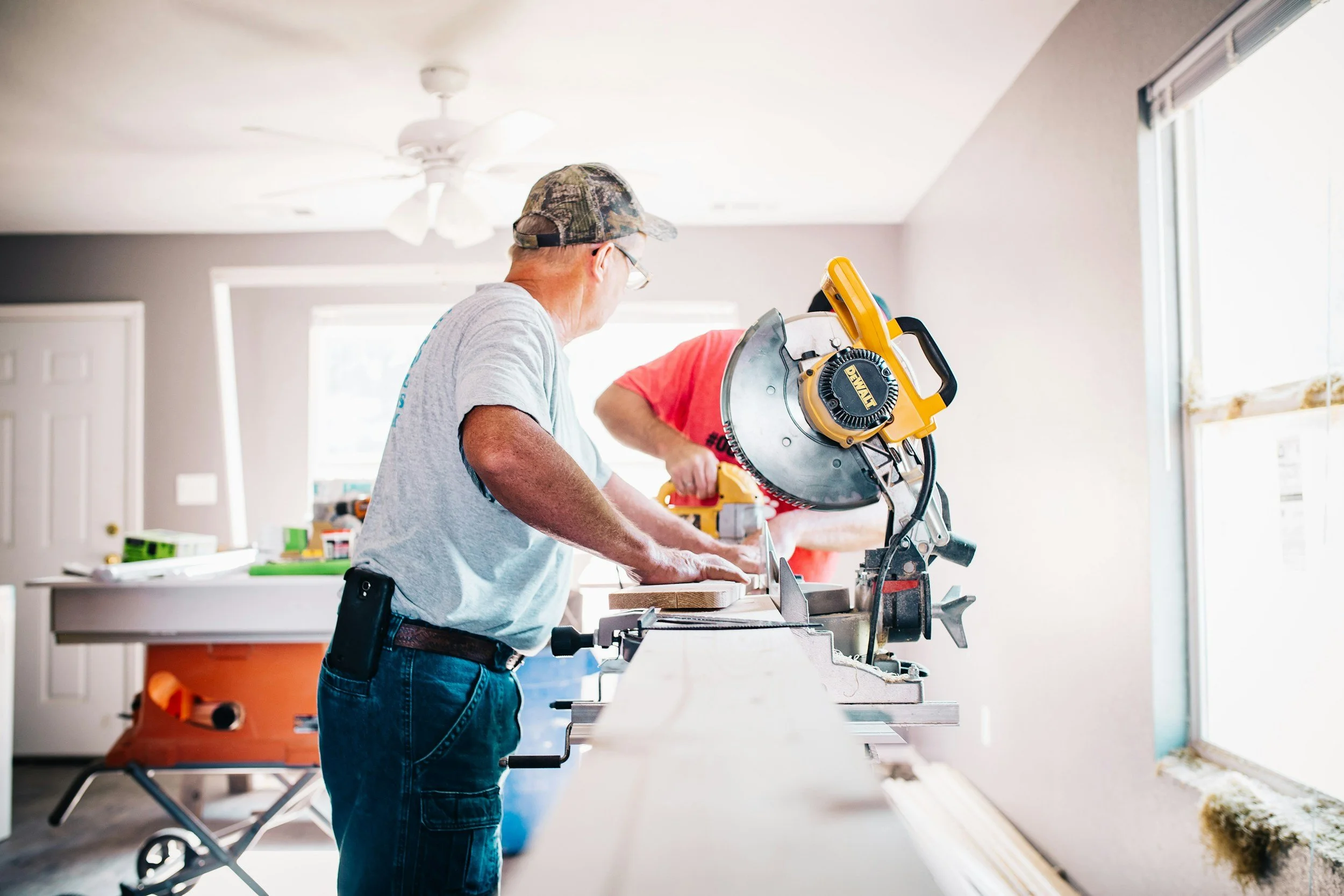 A man wearing glasses and a camouflage baseball cap uses a circular saw to cut wood in a bright room. Another person, dressed in a red shirt, assists nearby. The room appears to be under renovation with supplies on a table and an orange saw horse in the background.