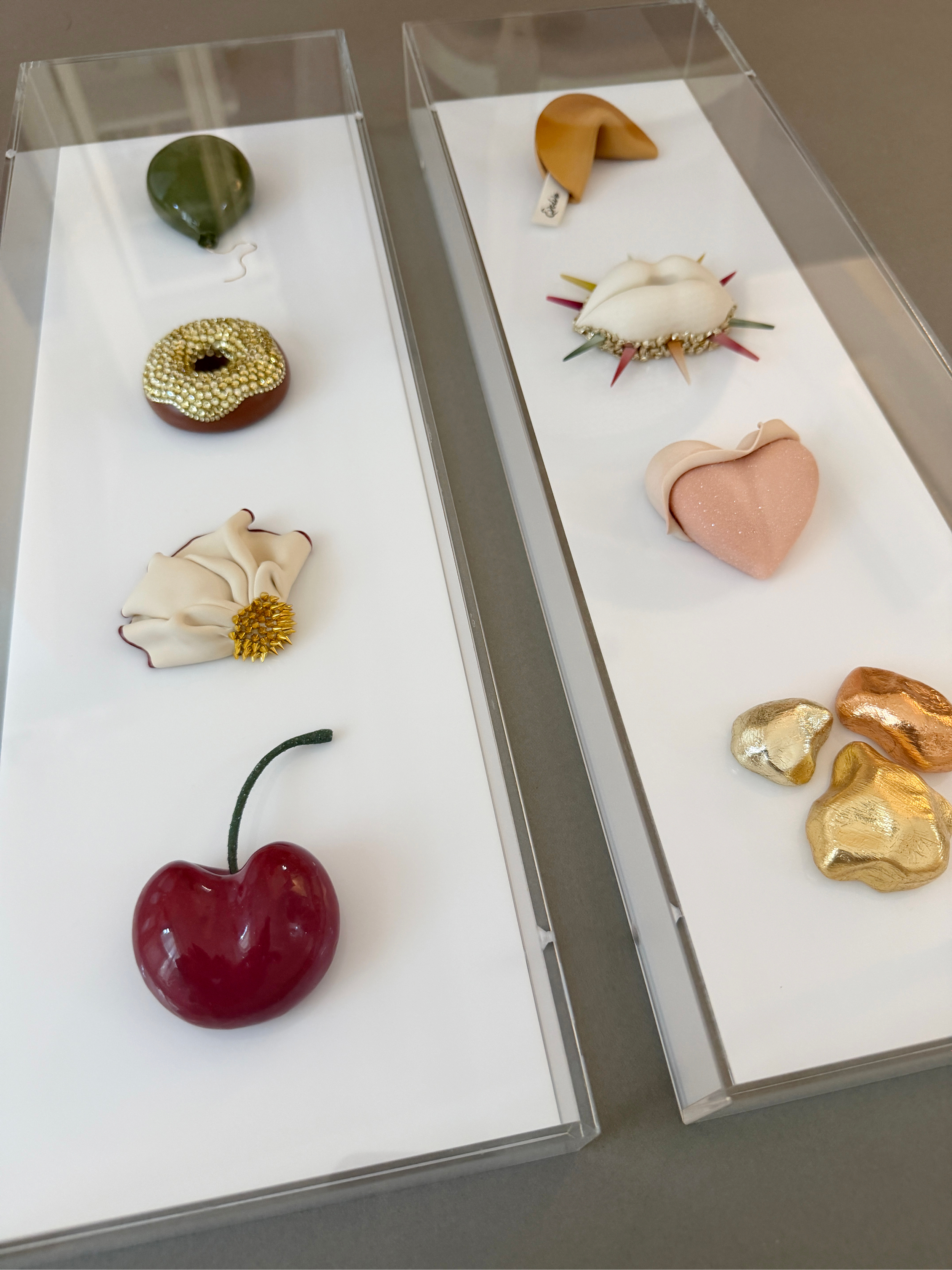 Display case with various decorative objects including a cherry, a beige heart, a white shell with colorful spikes, a beige crescent-shaped object, four gold-colored rock-like objects, a green shamrock-shaped object, a golden beaded donut, a gold spi