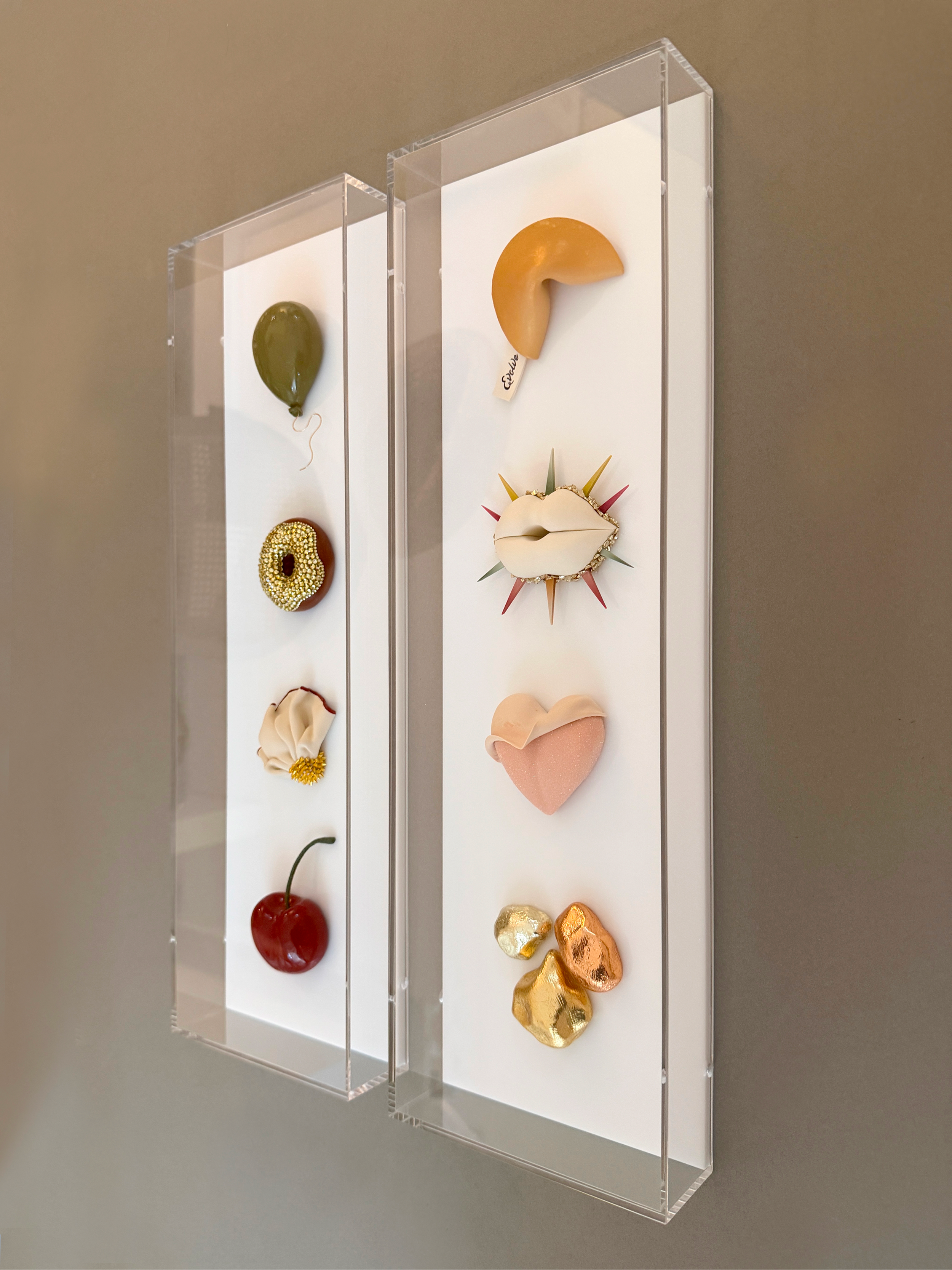 Art installation consisting of various objects resembling a balloon, a piece of meat with spikes, a cherry, and shiny gold objects, arranged in a display case.