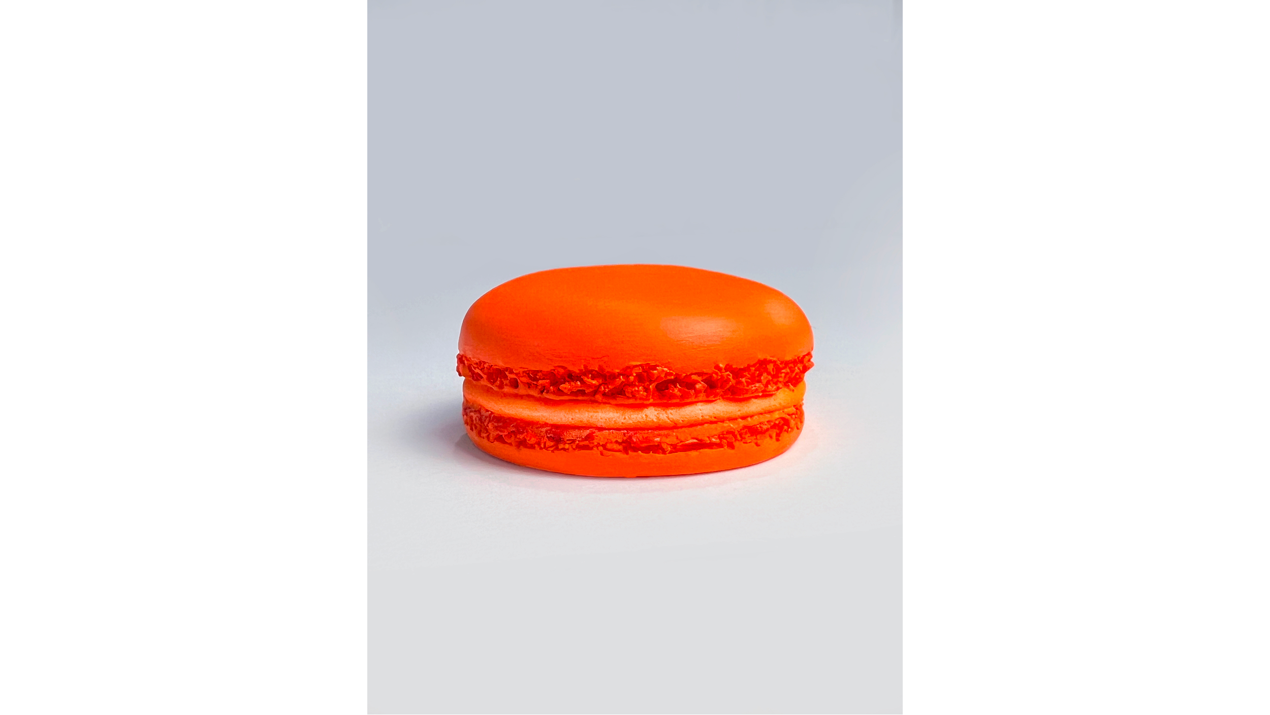 An orange macaron with a shiny top and textured filling, placed on a white surface with a plain gray background.