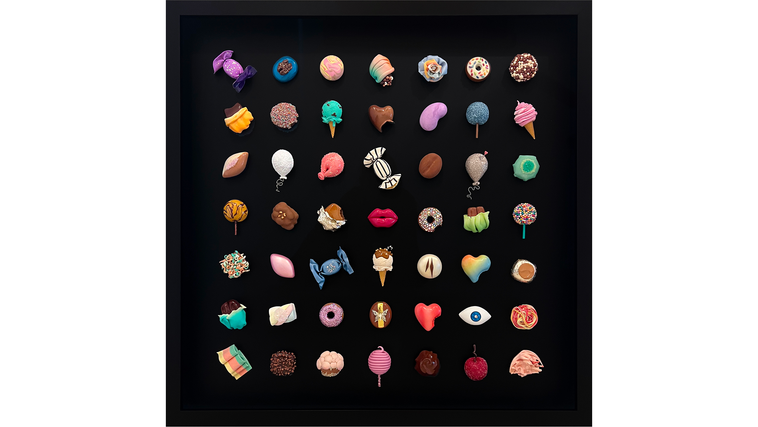 A display case with multiple colorful, decorative confections, including cupcakes, candies, and chocolates, arranged in rows on a black background.