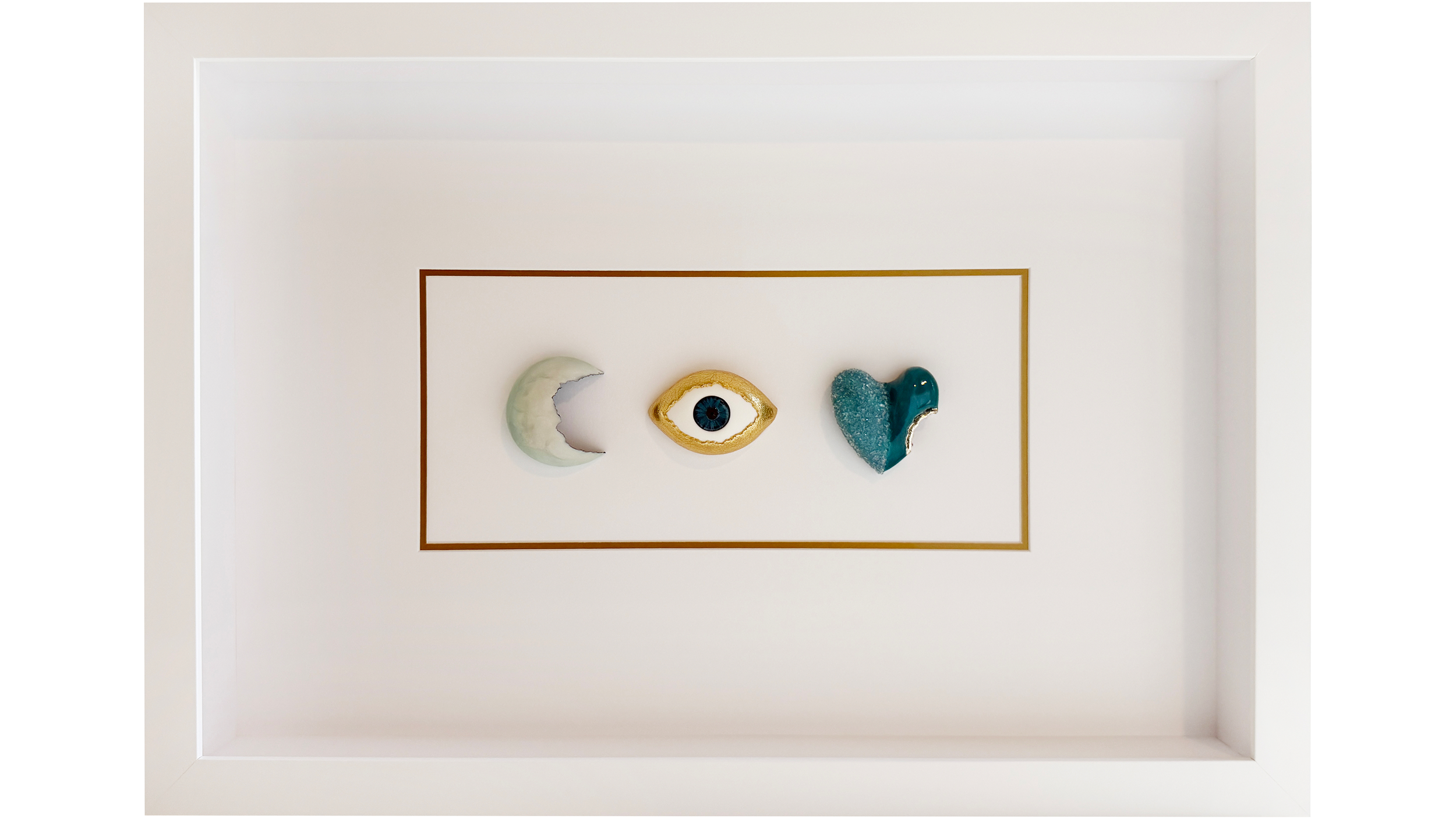 Decorative art piece with three ceramic elements: a crescent moon, an eye, and a heart, mounted on a white wall within a gold frame.