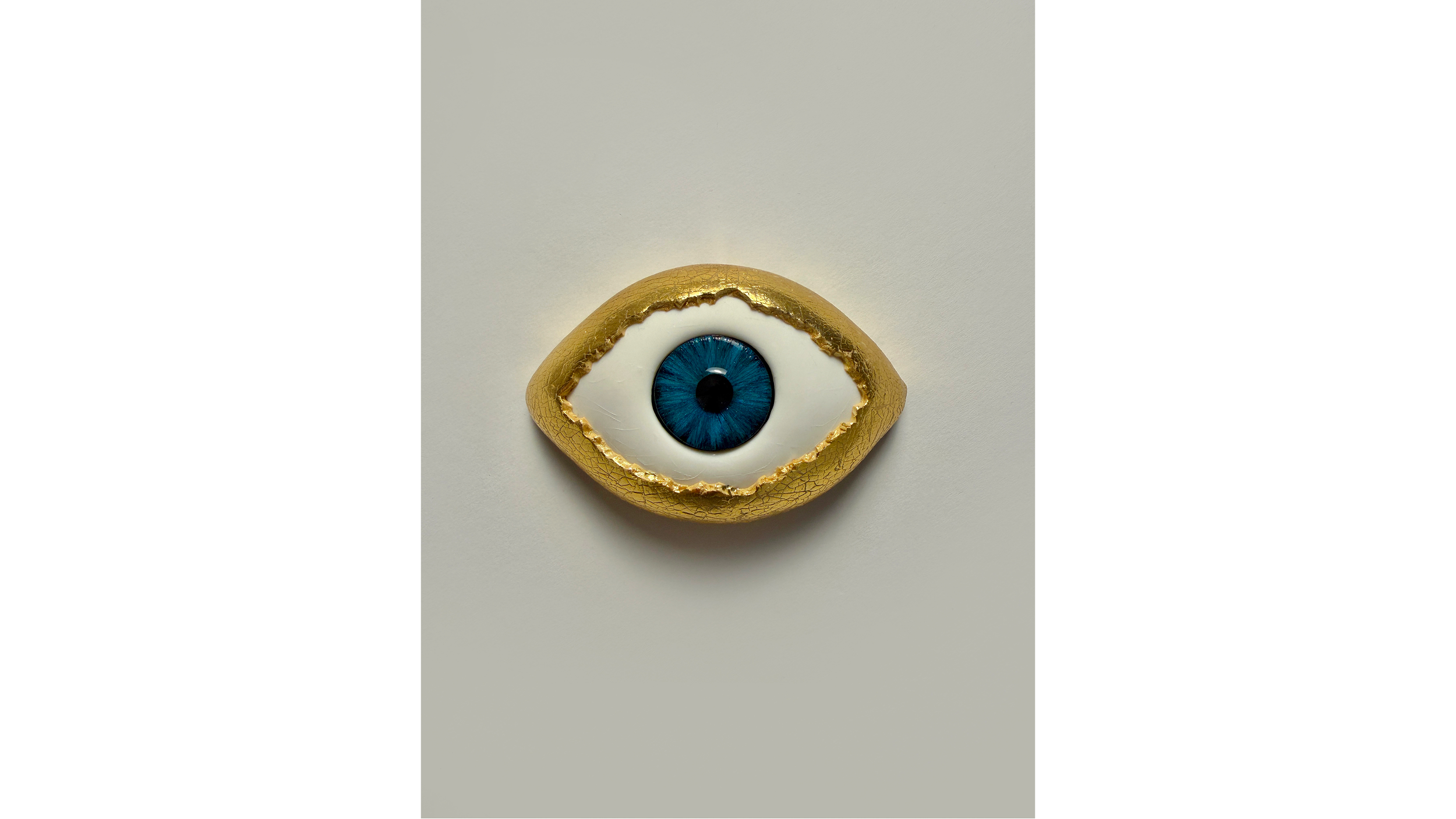 A decorative object resembling an eye, with a blue iris and a black pupil, surrounded by a white sclera, embedded in a gold-colored textured shape, on a plain, light background.