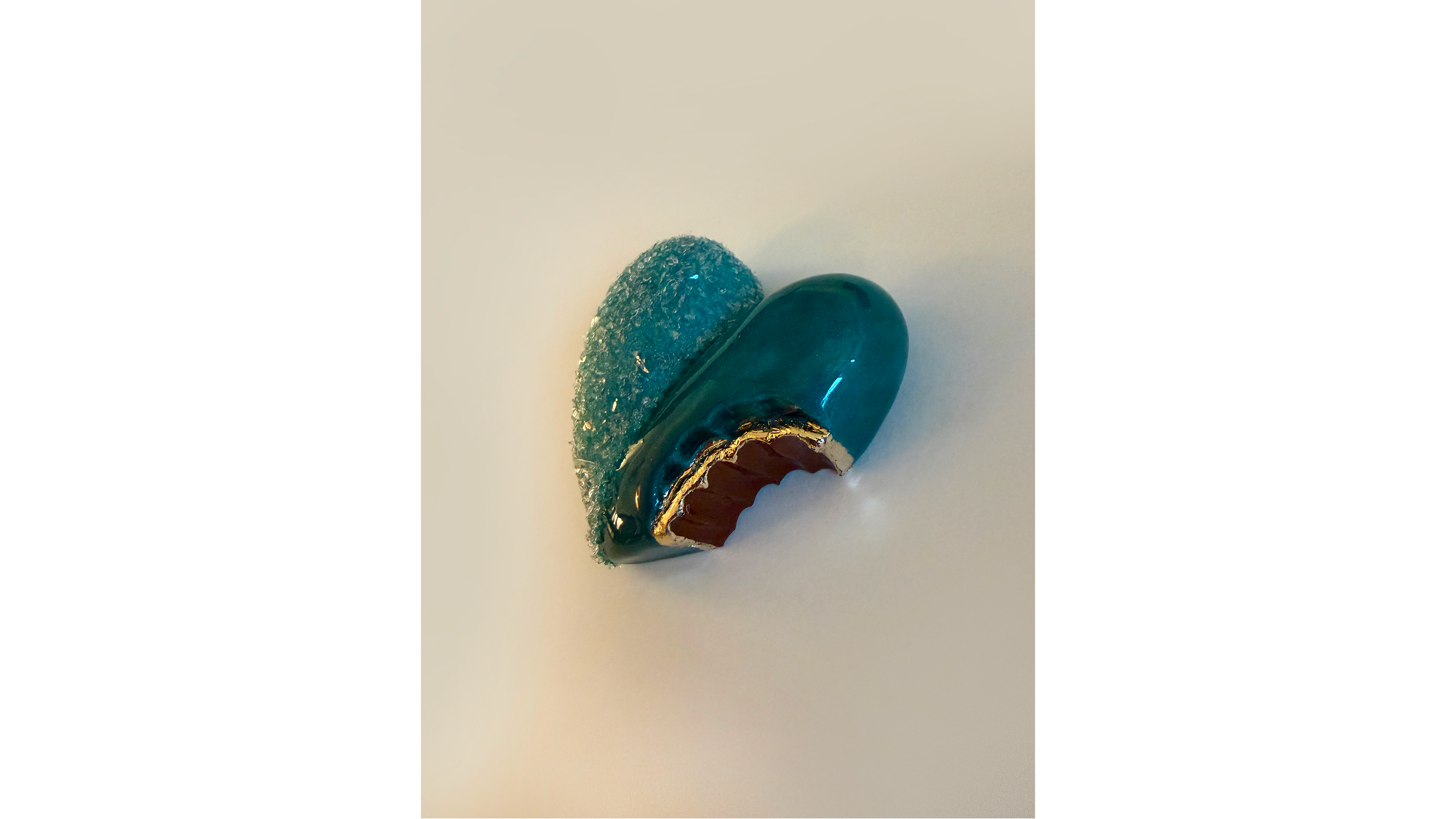 A decorative item resembling a fish, with a shiny teal body, a gold-accented mouth, and a textured, sparkly tail, placed on a white surface.
