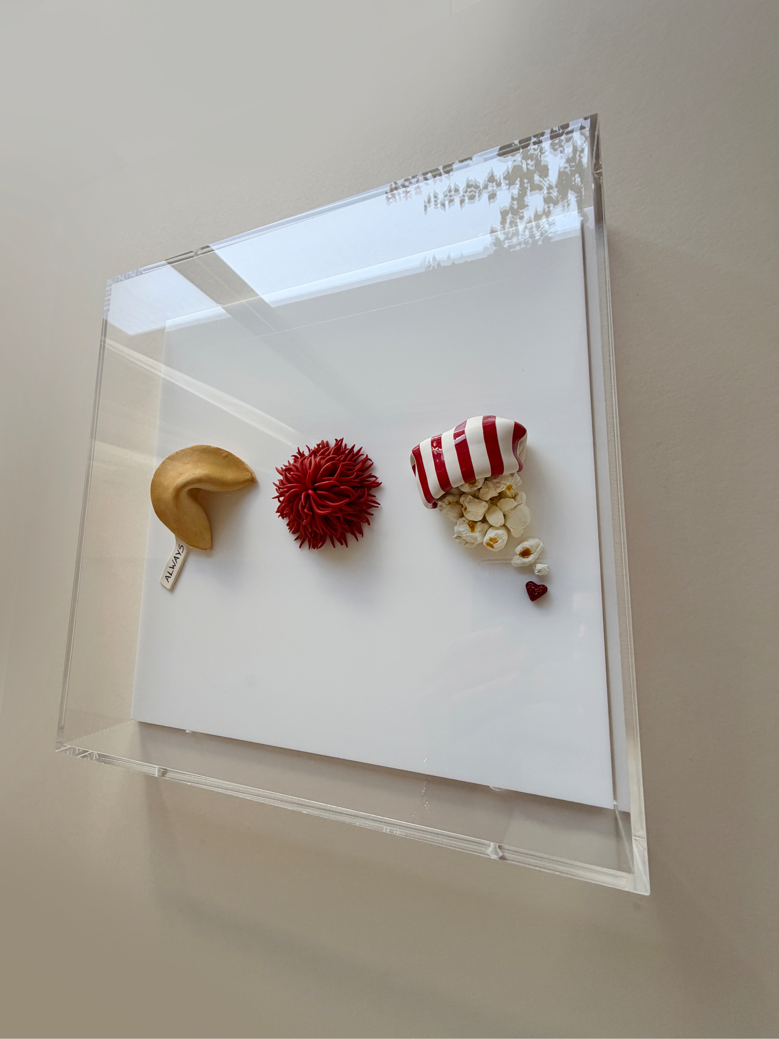 A clear acrylic display case with three small objects inside: a beige crescent moon with a small white tag reading 'ALWAYS,' a red spiky ball, and a piece of popcorn with a red and white striped wrapper, set against a white background.