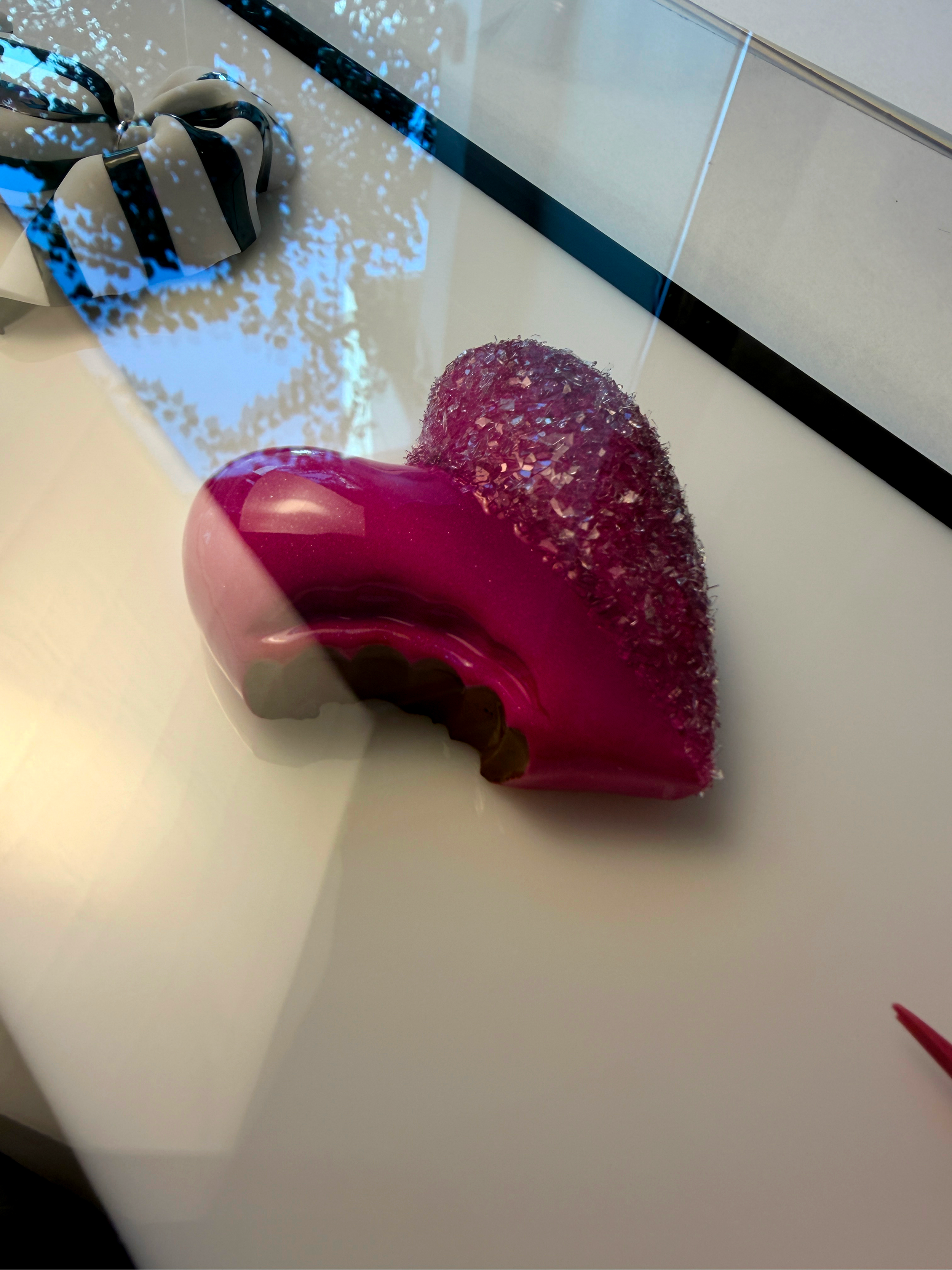A shiny, deep pink or magenta nail polish bottle with a rounded base and a sleek, curved shape, placed on a white surface. In the background, there is a glass mirror, reflecting a blurred outdoor scene with blue sky and tree branches, and a decorativ