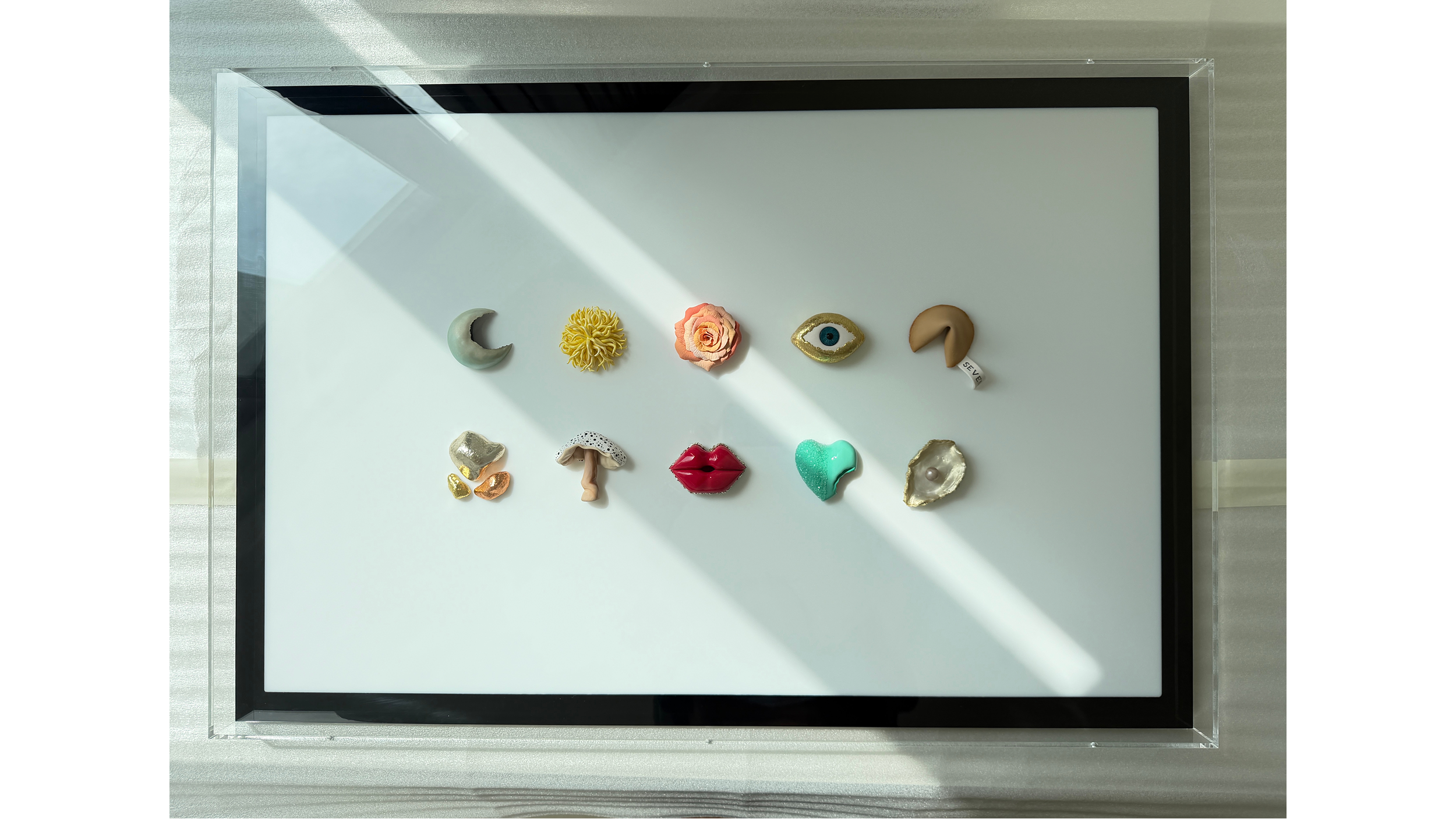 A framed artwork displaying an arrangement of various shaped objects, including a crescent moon, a yellow flower, a pink flower, an eye, a coil, golden and metallic stones, a mushroom, red lips, a heart, and a seashell, all on a white background with