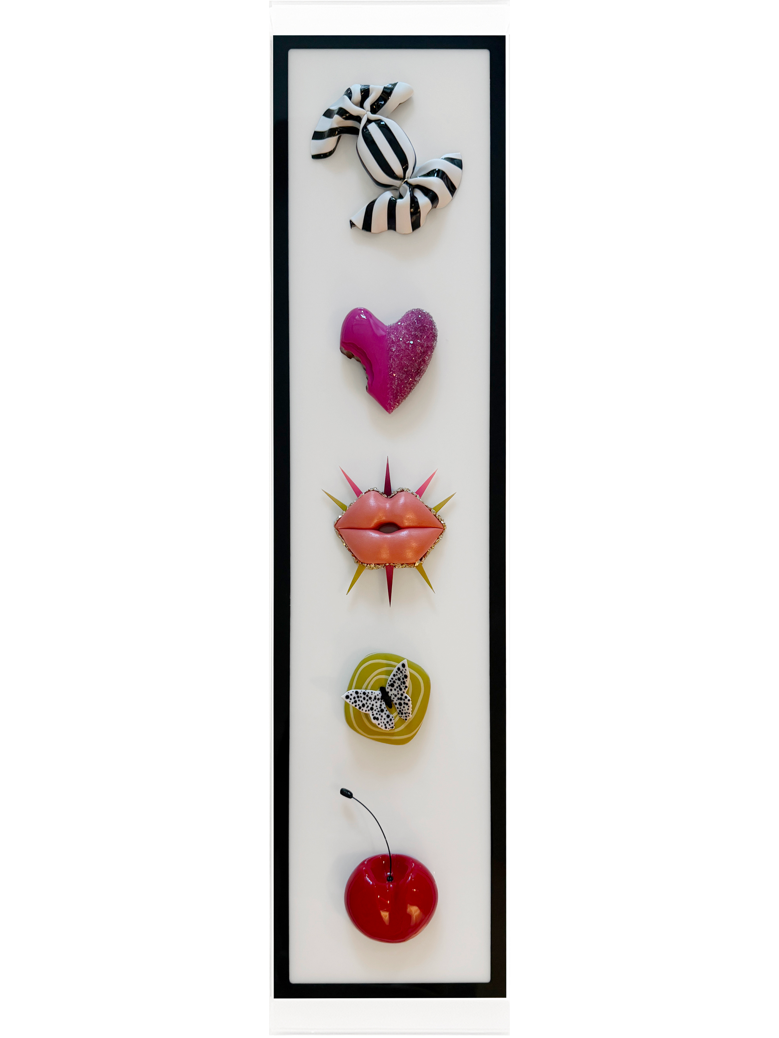 A framed wall art piece with decorative objects arranged vertically, including a black and white striped bow at the top, a pink heart-shaped object in the middle, and a red cherry with a green stem at the bottom.