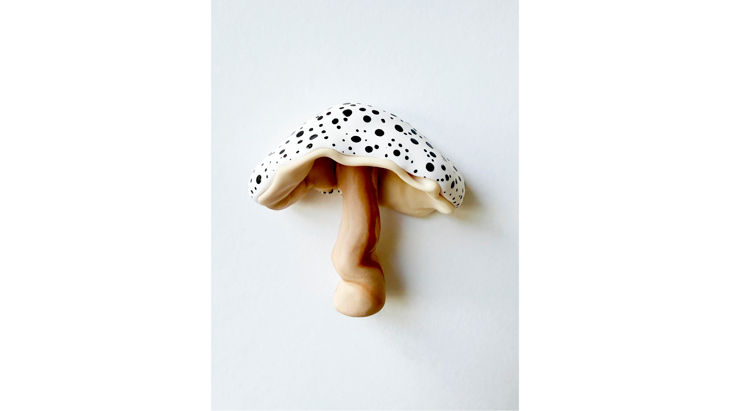 A realistic sculpture of a mushroom with a white cap dotted with black spots, and a beige stem, held by a human hand against a plain white background.