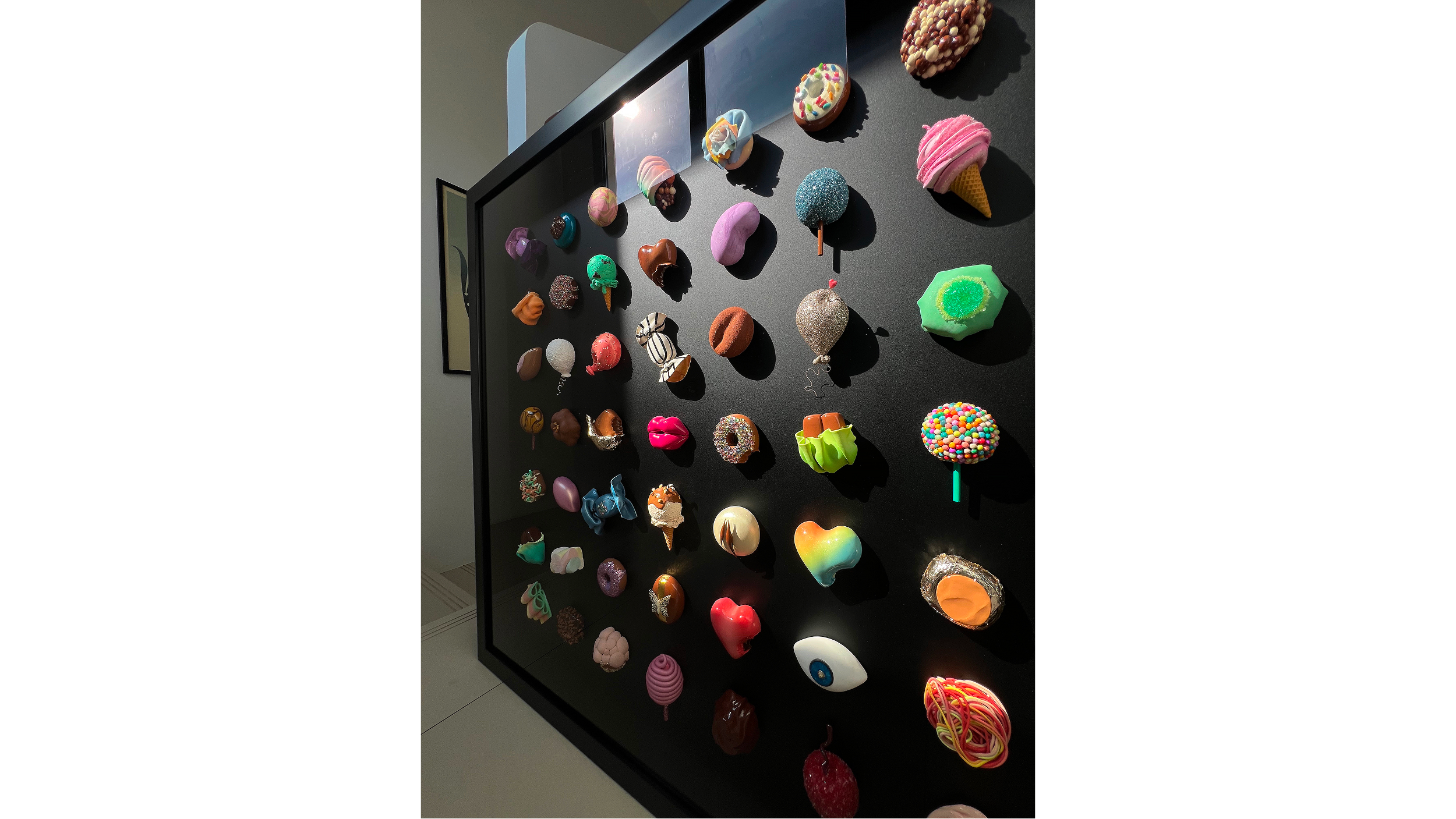 Display of various colorful, decorative objects, possibly handmade crafts or art pieces, on a black table with a glass display case in the background.