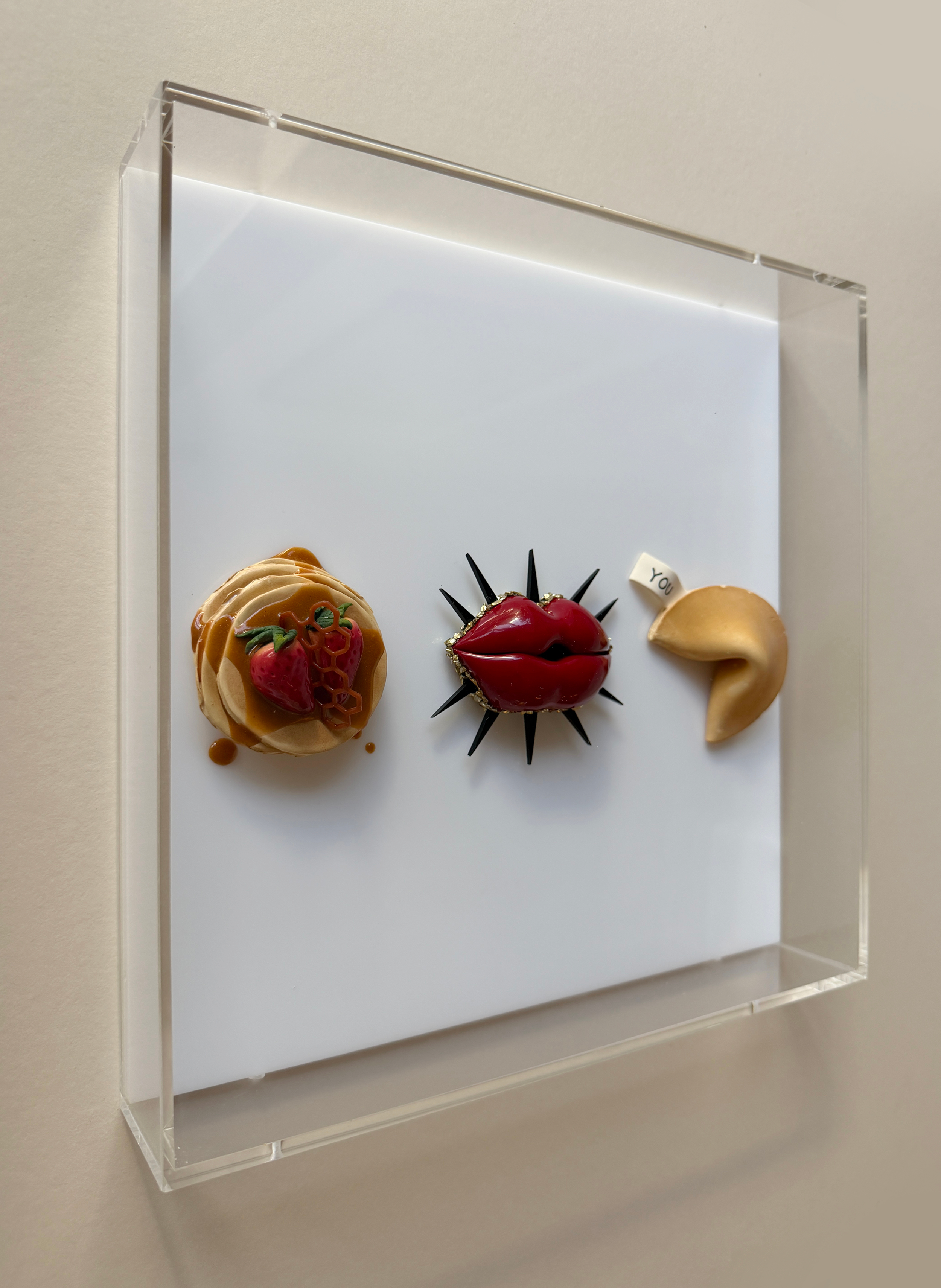 A white shadow box contains three decorative pastries, including a caramel pastry with berries, a glossy red lip-shaped pastry, and a golden crescent moon pastry, all with small tags on a white background.