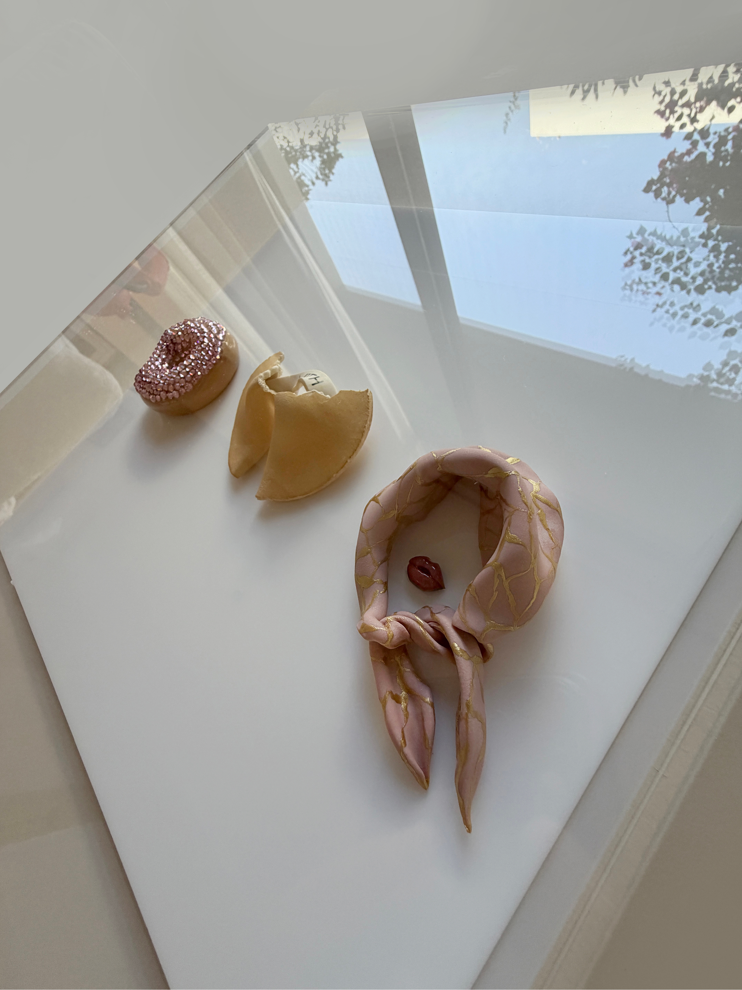 Decorative doughnuts, a crescent-shaped pastry, and a scarf with pink and gold design on a white surface near a window with trees outside.