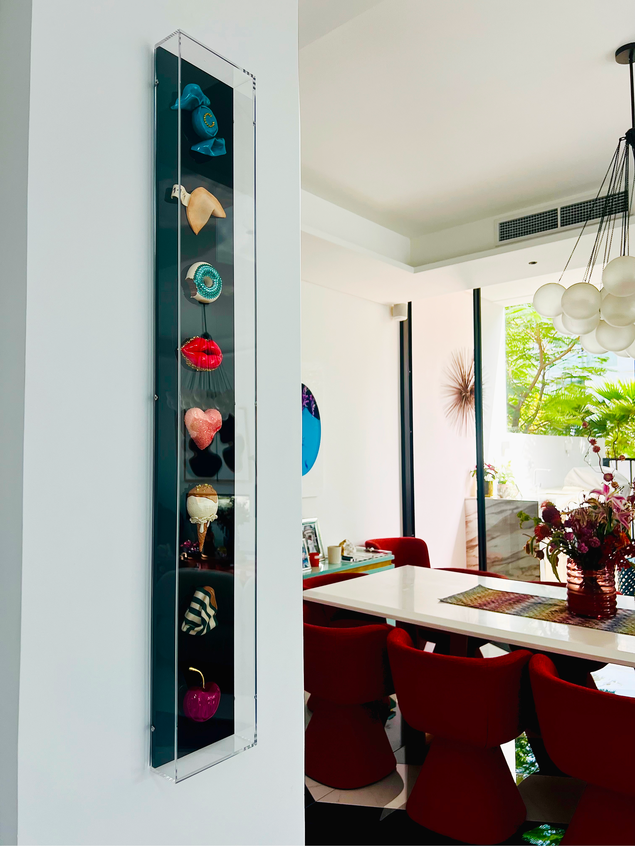 Interior view of a modern dining room with a white table, red velvet chairs, and a large window showing greenery outside. A contemporary chandelier hangs above the table, and a vase with flowers is on the table. A painted wall art and a display cabin