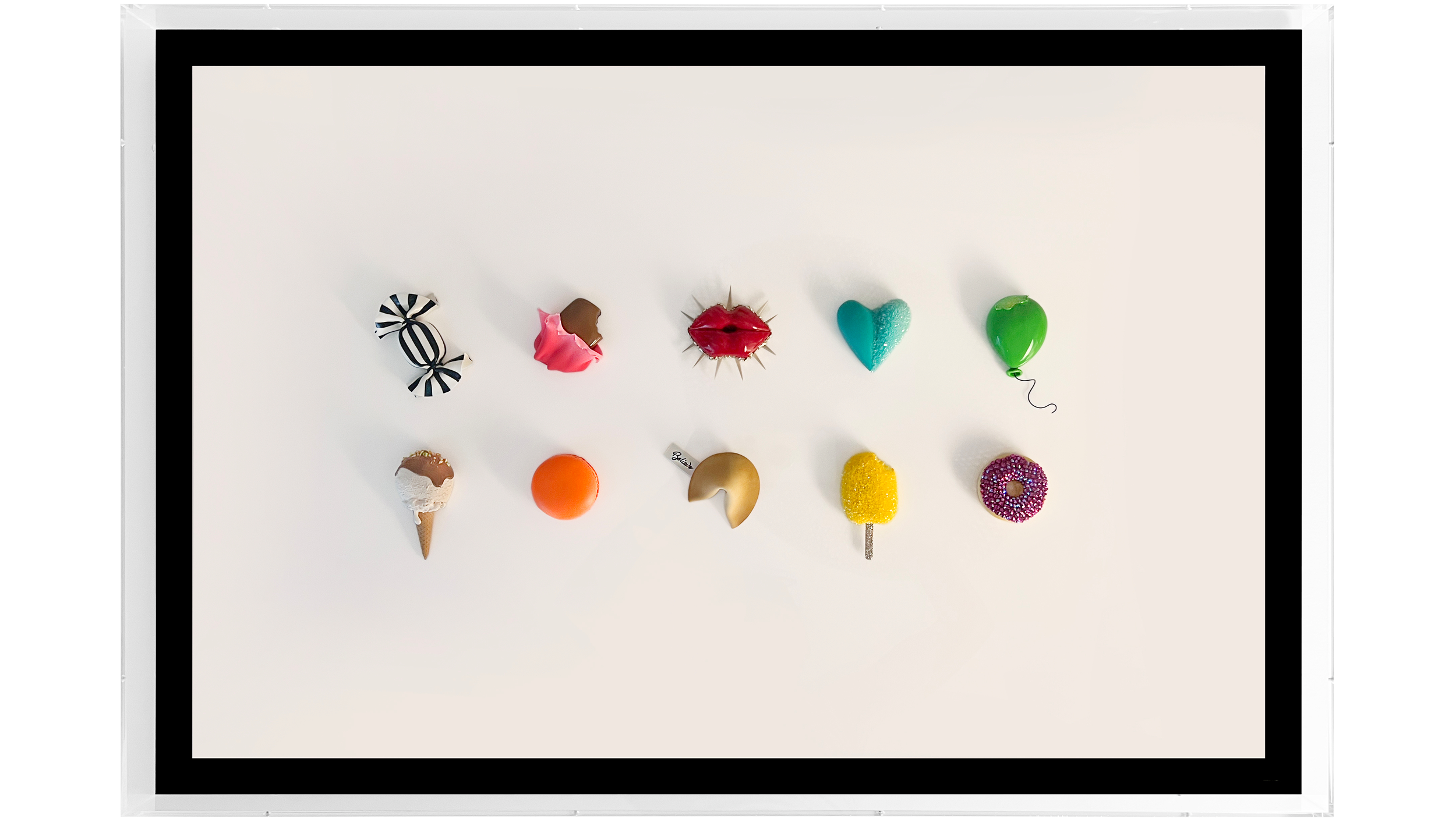 A framed display of decorative items arranged in two rows. The items include an ice cream cone, a round orange object, a crescent-shaped object, a yellow square, a donut with pink and white sprinkles, a wrapped candy, a pink rose decoration, a red li
