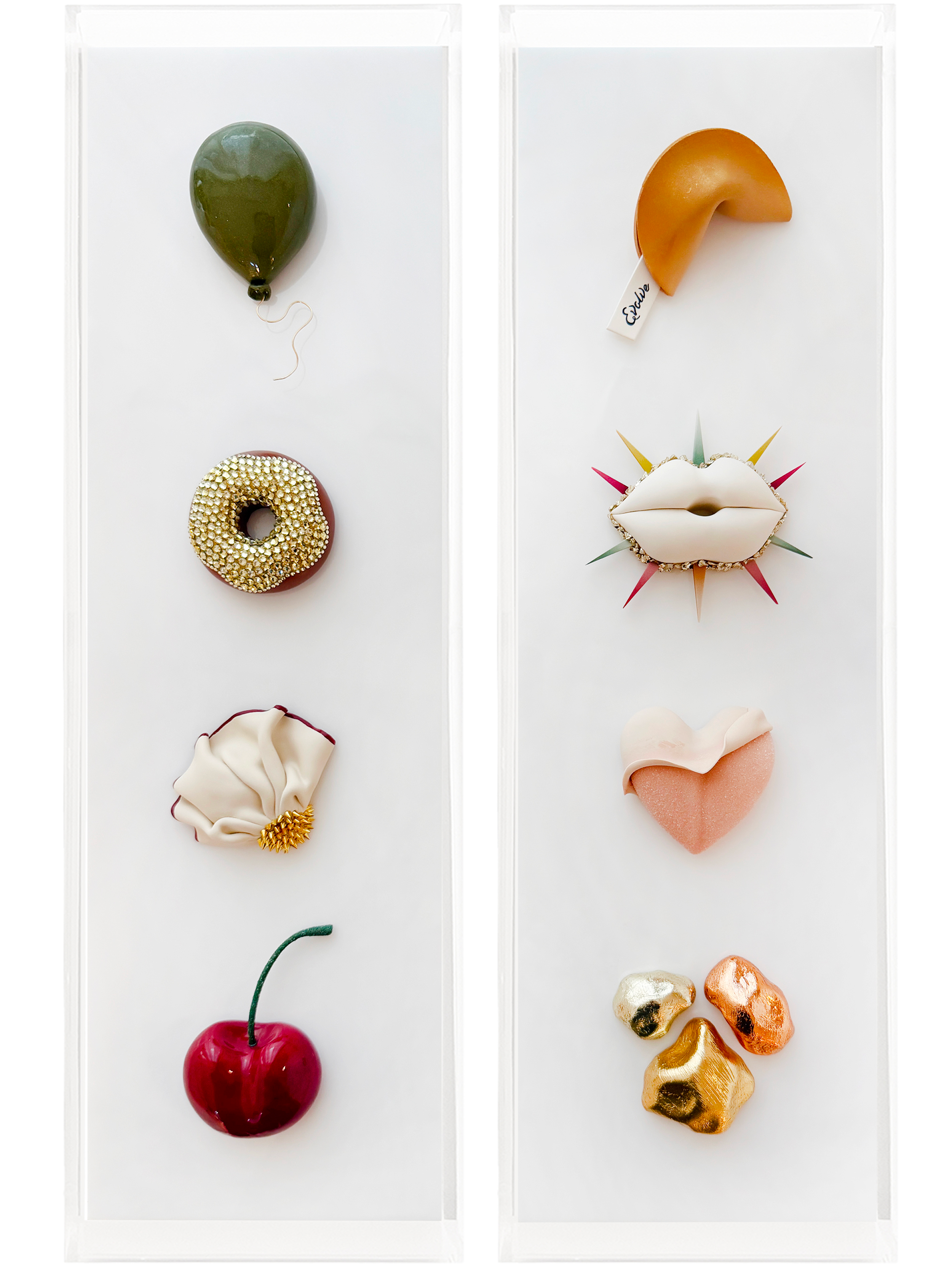 Two display cases with various decorative objects, including a green balloon, a donut with gold beads, a peach-colored crescent, a mouth-shaped object with colorful spikes, a pink heart-shaped item, and several gold and metallic rocks.