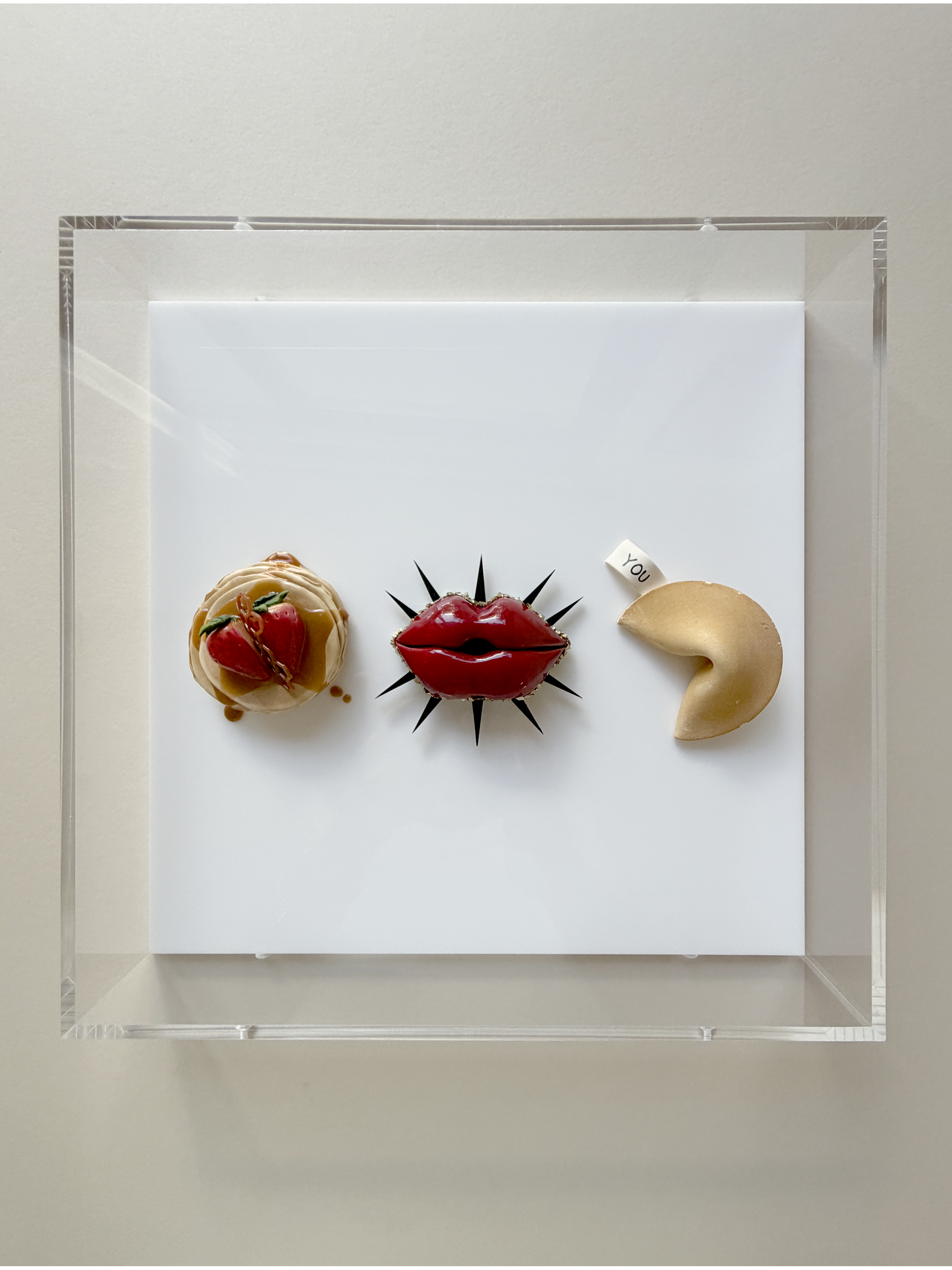 Decorative arrangement of three desserts on a white surface, with a white backdrop. The left dessert is a round pastry topped with strawberries and caramel drizzle. The middle dessert resembles a red lips with a spiky black decoration behind it. The 