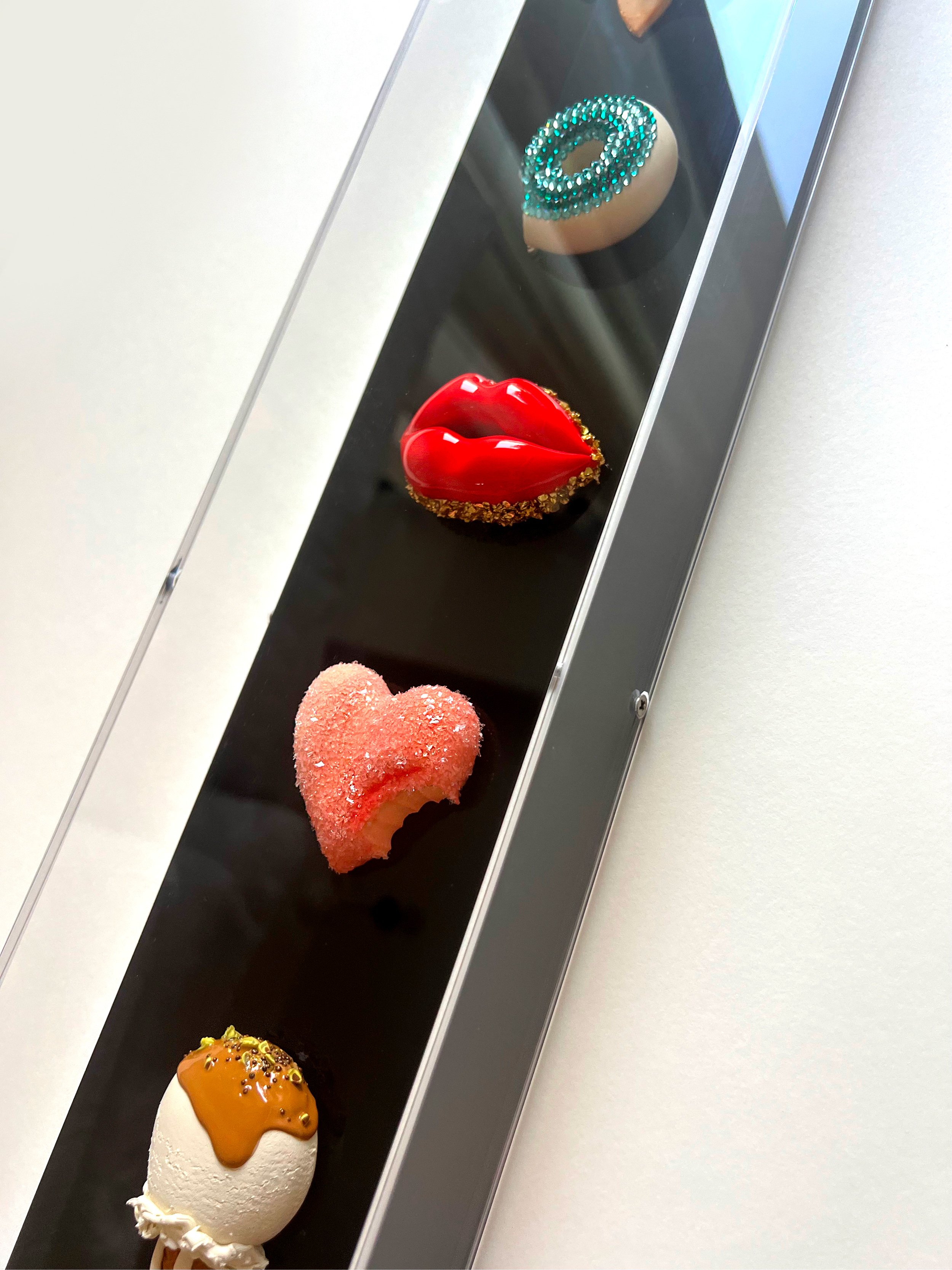 A display of four decorated desserts on a black tray. From top to bottom, a ring of teal rhinestones, a pair of red lips with gold accents and a glittery gold border, a pink heart-shaped cake with pink icing and sugar crystals, and a caramel-colored 